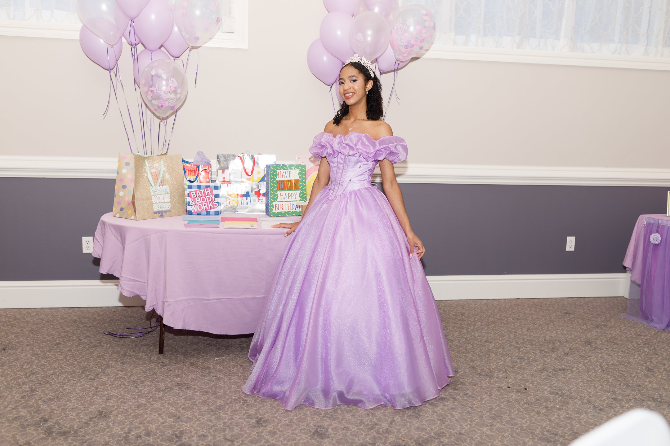 Young woman dressed in a purple princess gown and crown, standing at a birthday party table with pink balloons and gift bags.