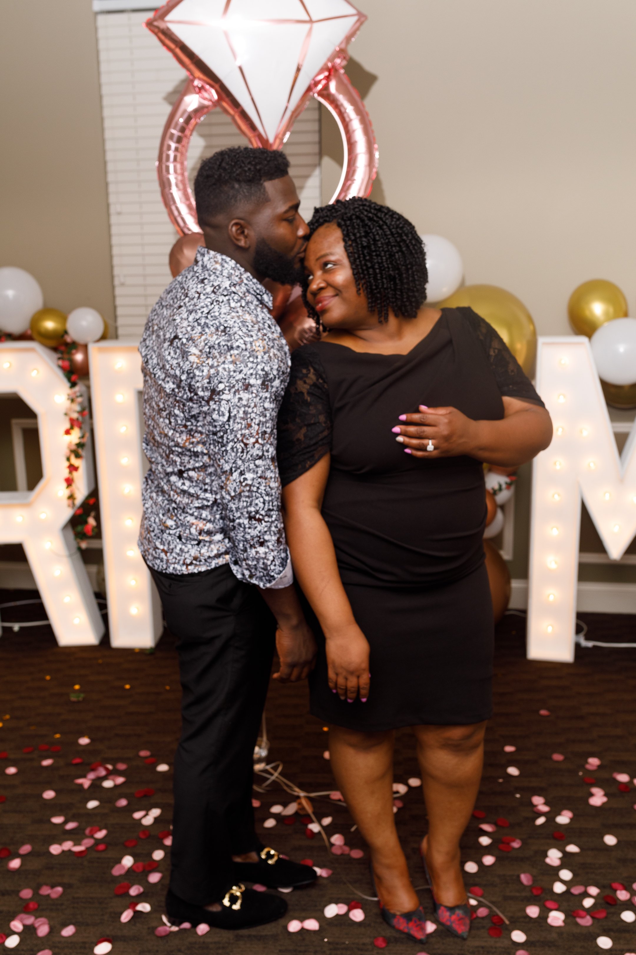 A couple standing close together at a celebration, with the man kissing the woman's cheek and the woman smiling. Behind them are large illuminated letters spelling 'LOVE,' balloons, and decorations including a diamond-shaped balloon and rose gold bal