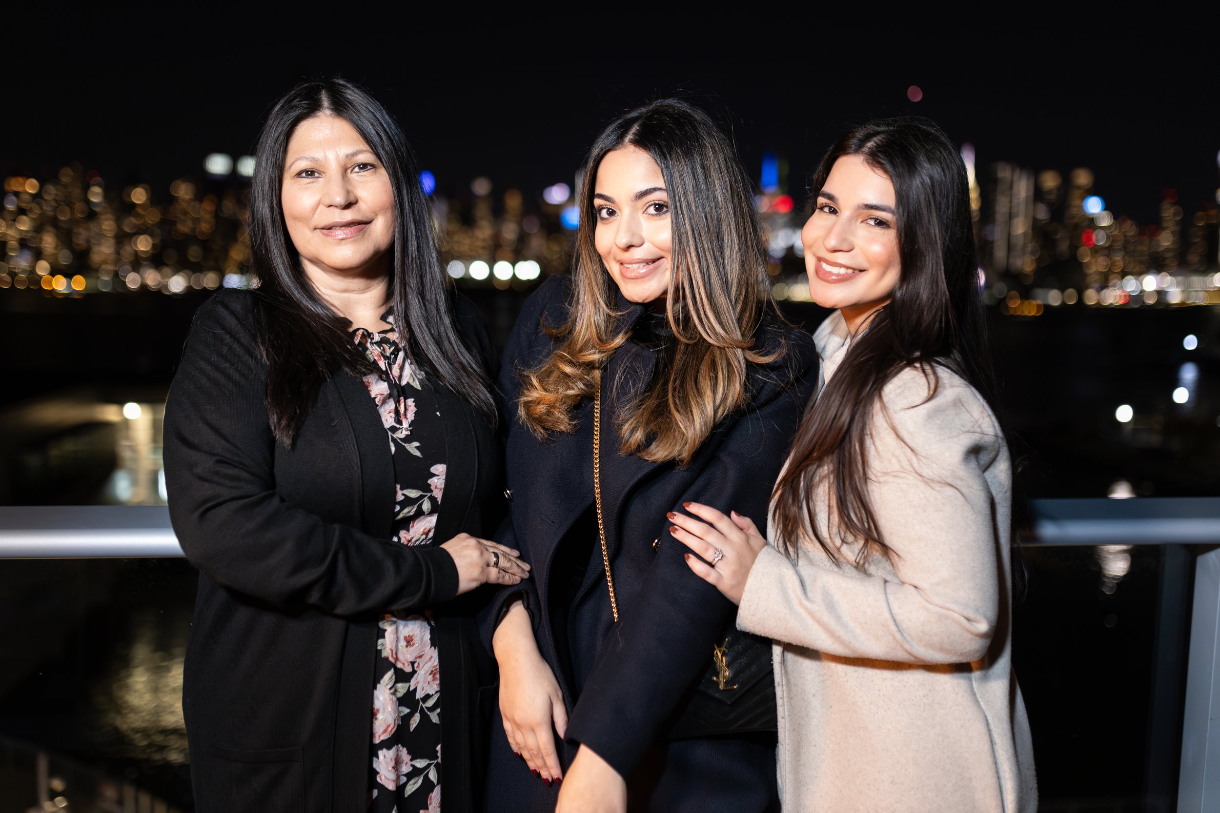 Three women standing on a balcony at night with city lights in the background, smiling and posing for the photo.