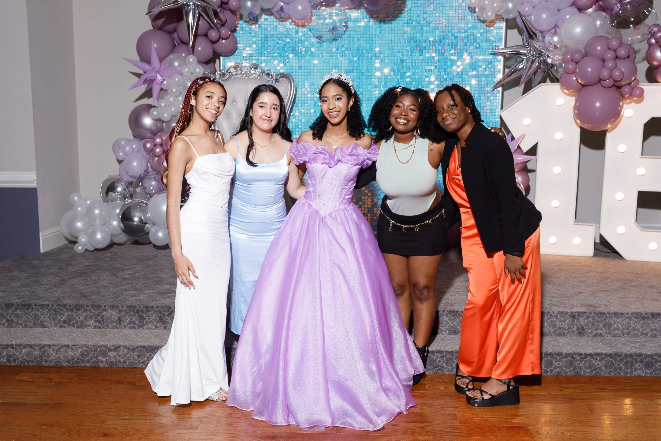 Five women dressed in formal and colorful outfits standing together at a celebration, with balloons and decorative backdrop.