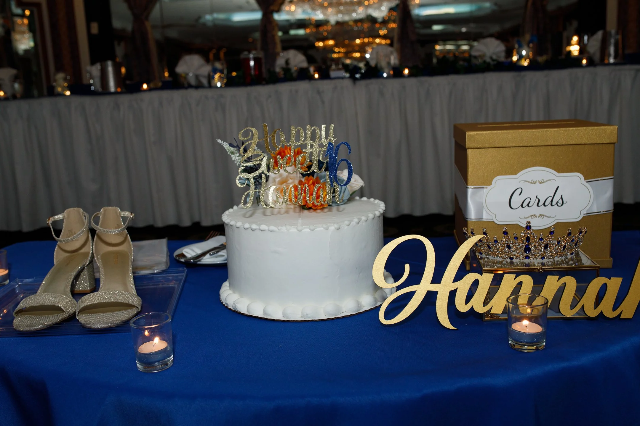 A decorated table with a white cake topped with a "happy 15th birthday Hannah" cake topper, gold high-heeled shoes, a "Cards" box, a floral crown, a large gold "Hannah" sign, and tea light candles, set in a banquet hall.