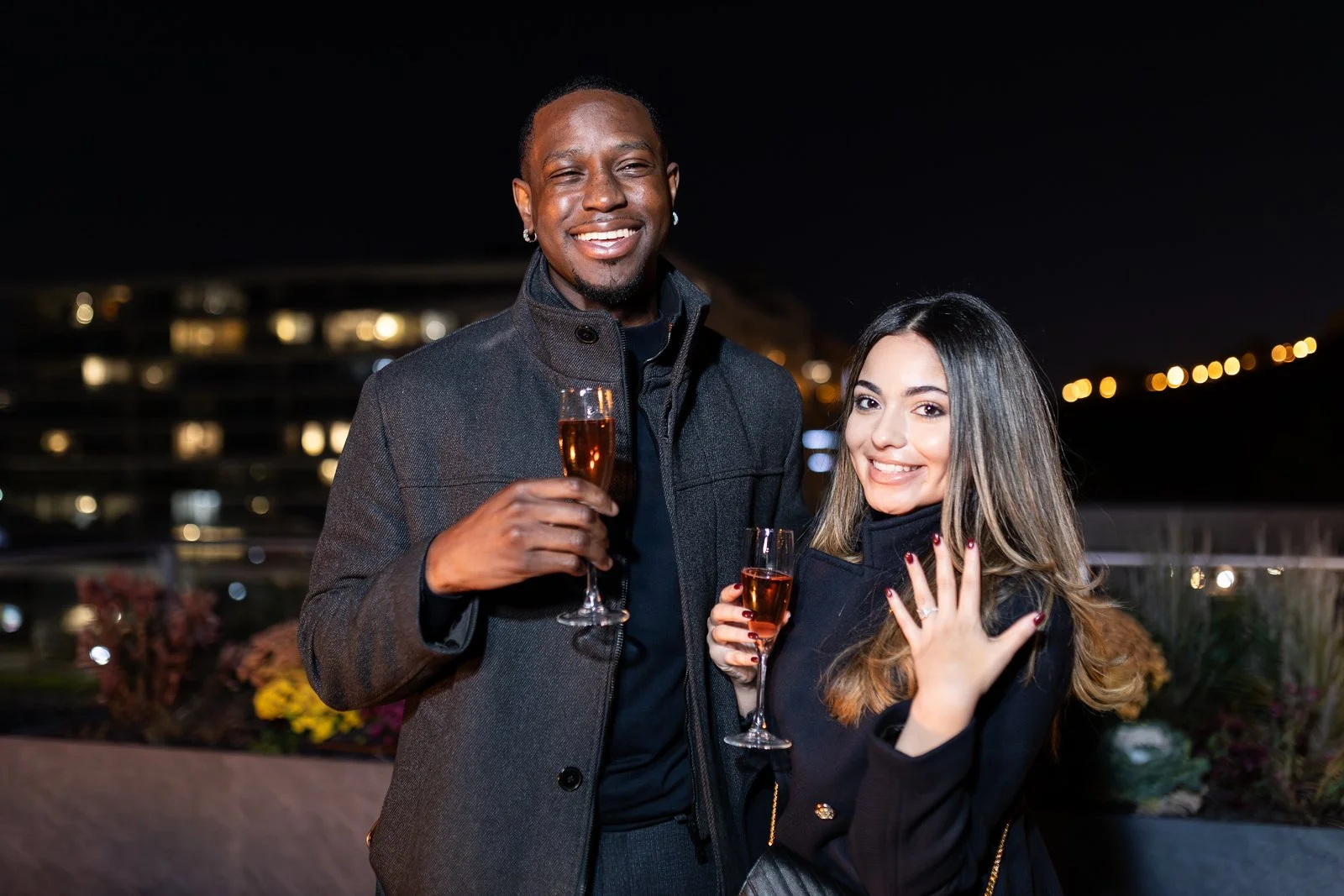 A man and woman holding glasses of rosé champagne, smiling at an outdoor nighttime event with city lights in the background.