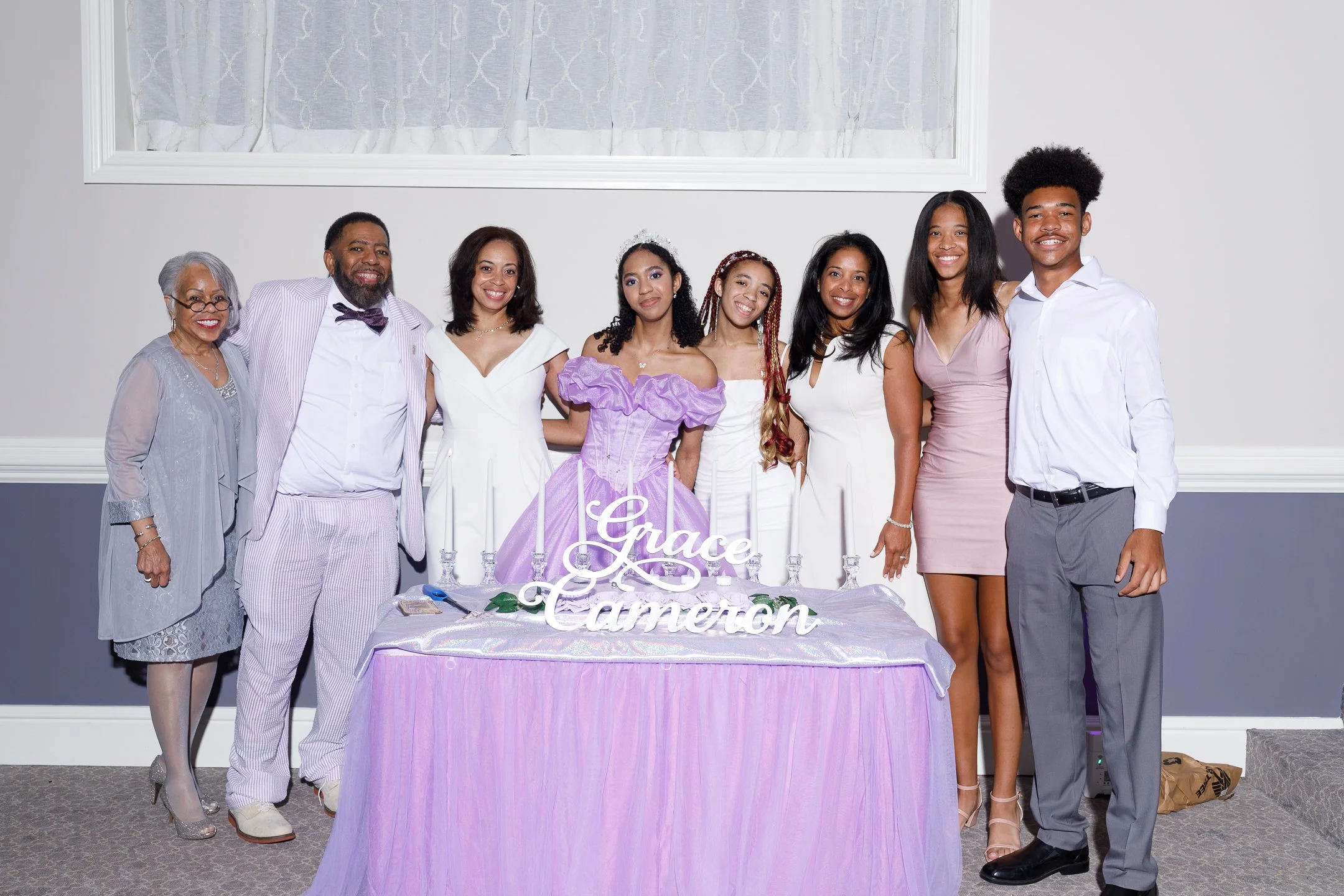 Group of women and men at a celebration, standing behind a table with a sign that says 'Grace Cameron', dressed in formal and semi-formal attire.