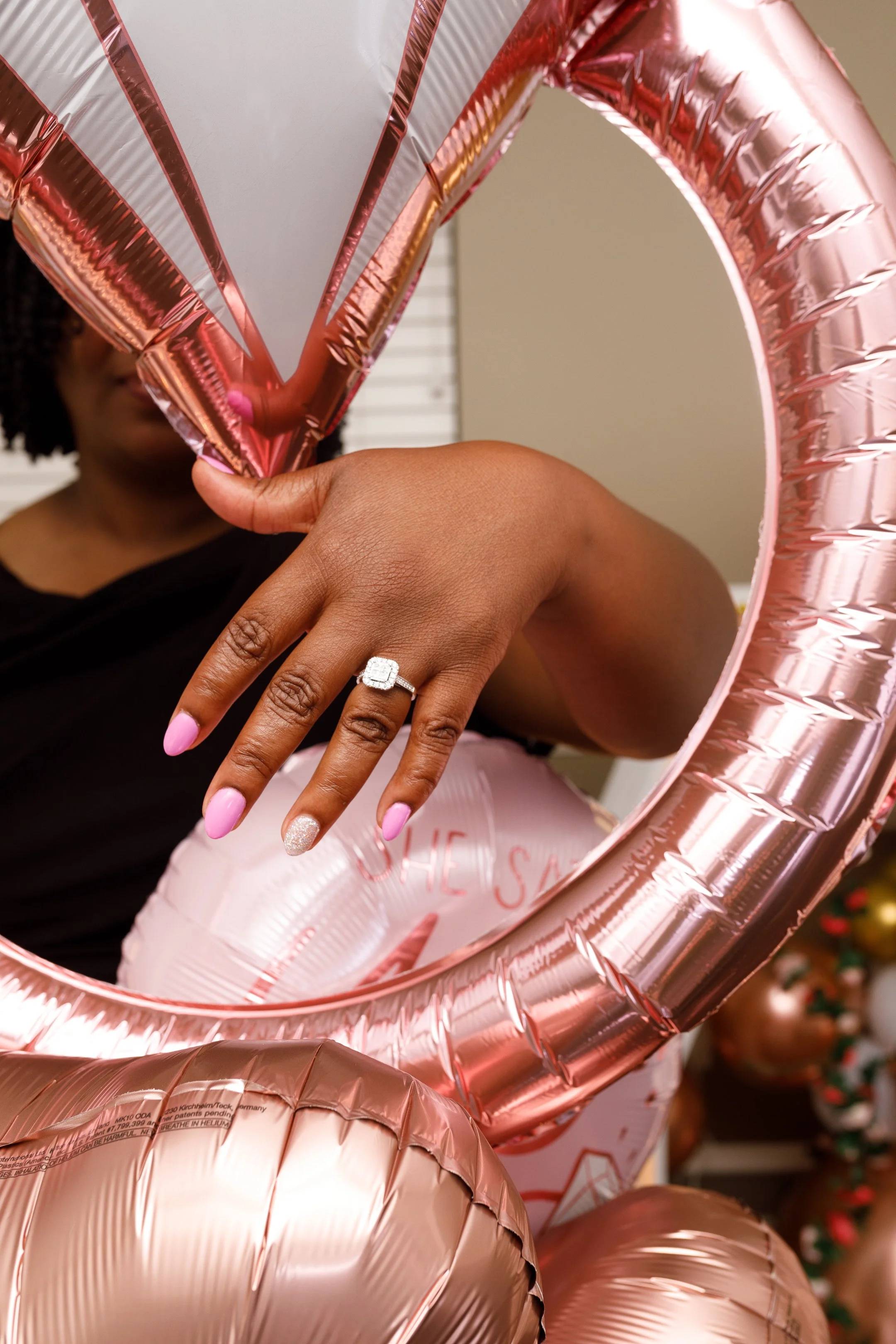 A woman shows off her engagement ring through a heart-shaped pink foil balloon, with additional pink balloons in the background.