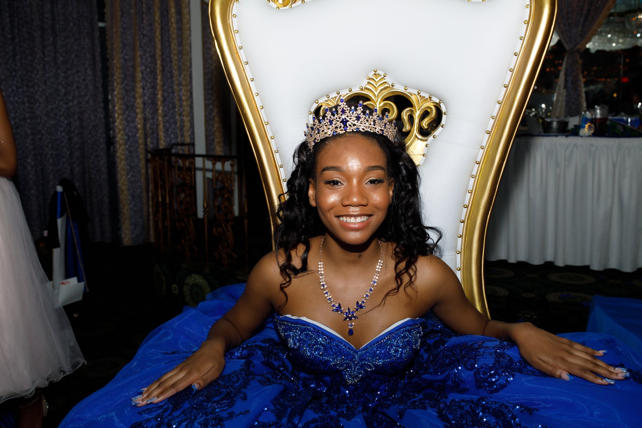 Young woman smiling, wearing a blue gown and a silver tiara, seated on a throne with a white and gold backrest at a celebration event.