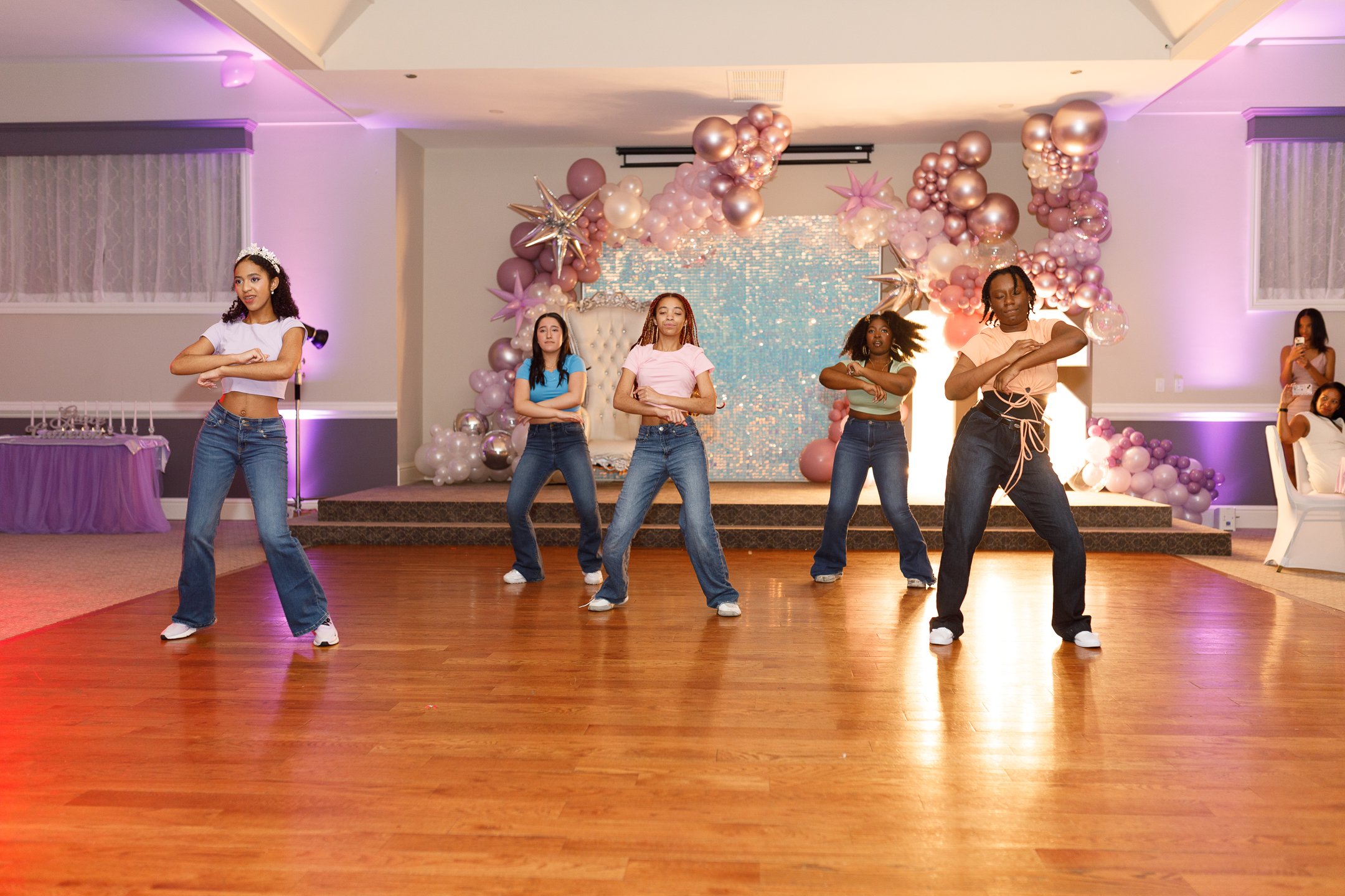 Five girls performing a dance routine on a wooden floor stage decorated with pink and purple balloons and star-shaped ornaments, with an audience watching in the background.