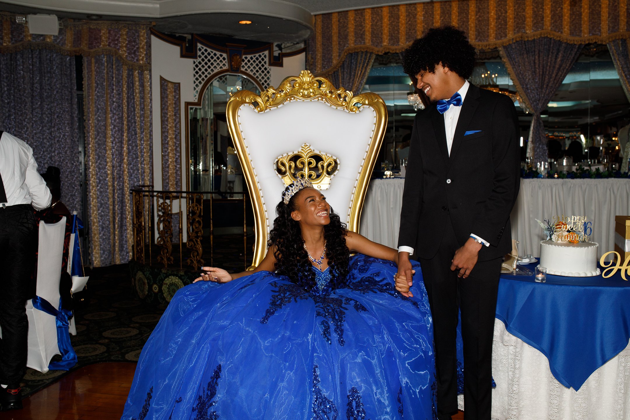 A woman in a royal blue ball gown and tiara sitting on a white and gold throne, smiling up at a man in a black tuxedo with a blue bow tie, during a celebration in a decorated banquet hall.