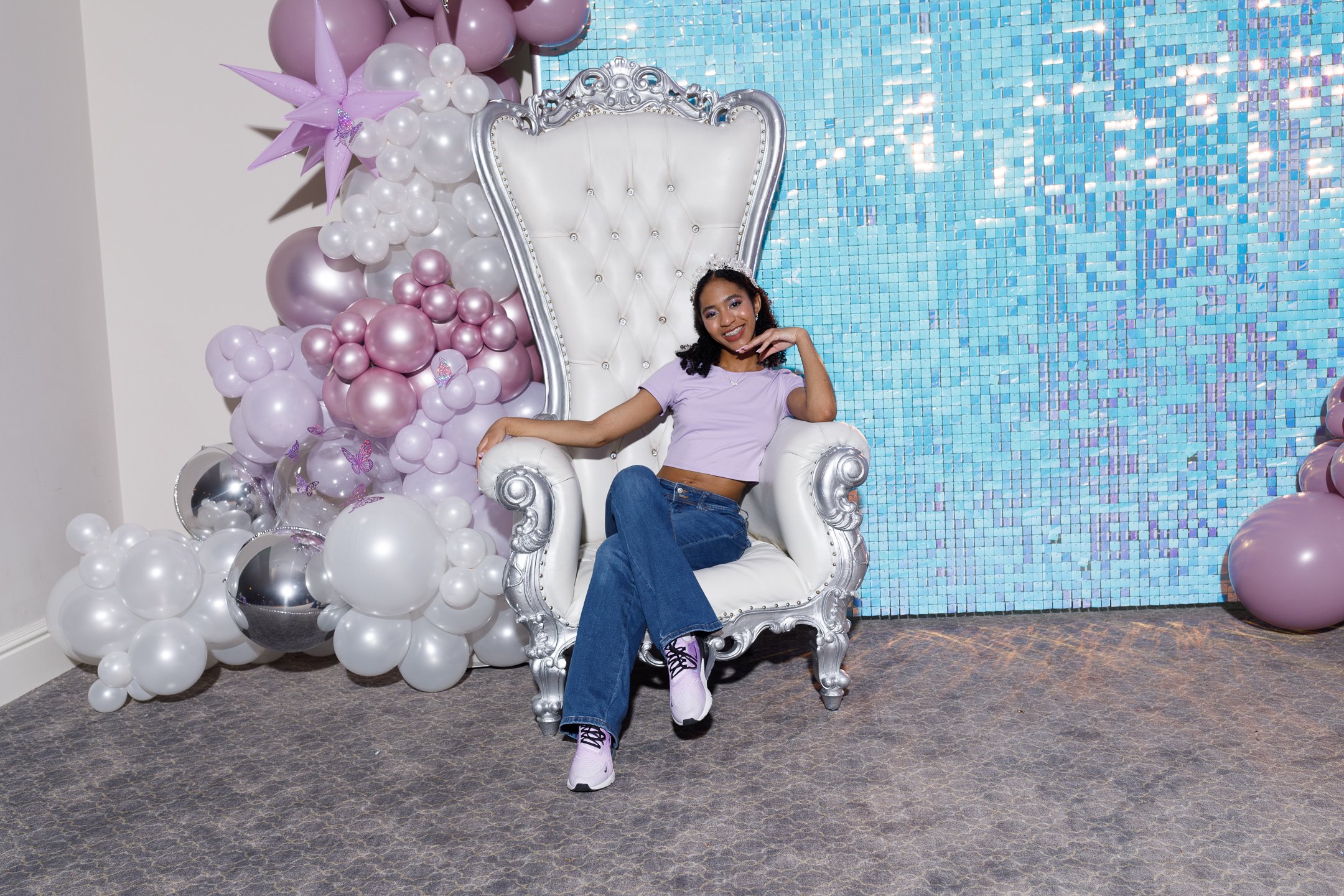 Young woman sitting on a large ornate white chair with silver accents, smiling, wearing a lavender crop top, blue jeans, and sneakers, with a backdrop of pink, white, and silver balloons and a blue mosaic wall.