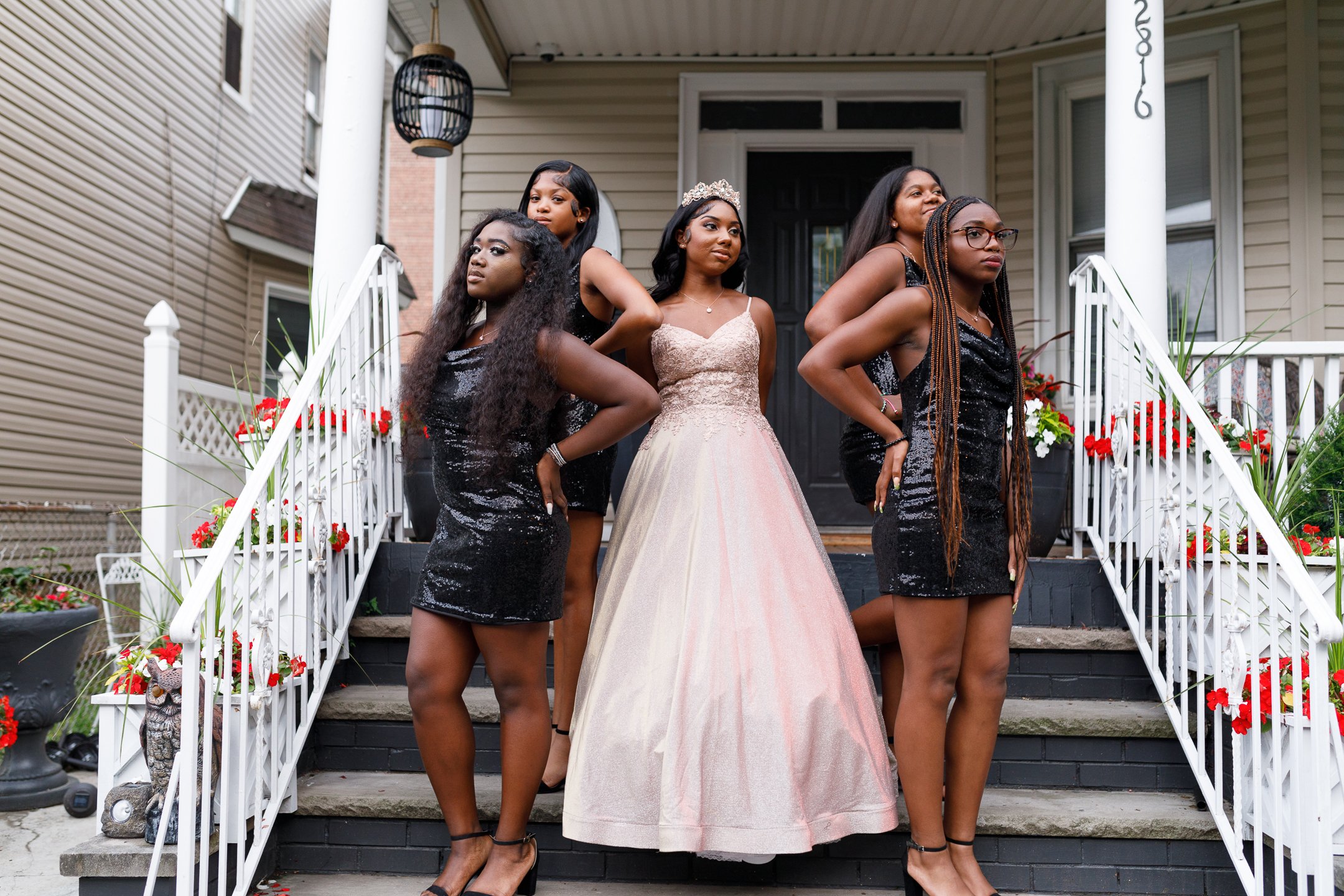 Group of five women standing on front porch steps, with three women in black dresses and a woman in a light-colored gown, posing confidently for a photo.