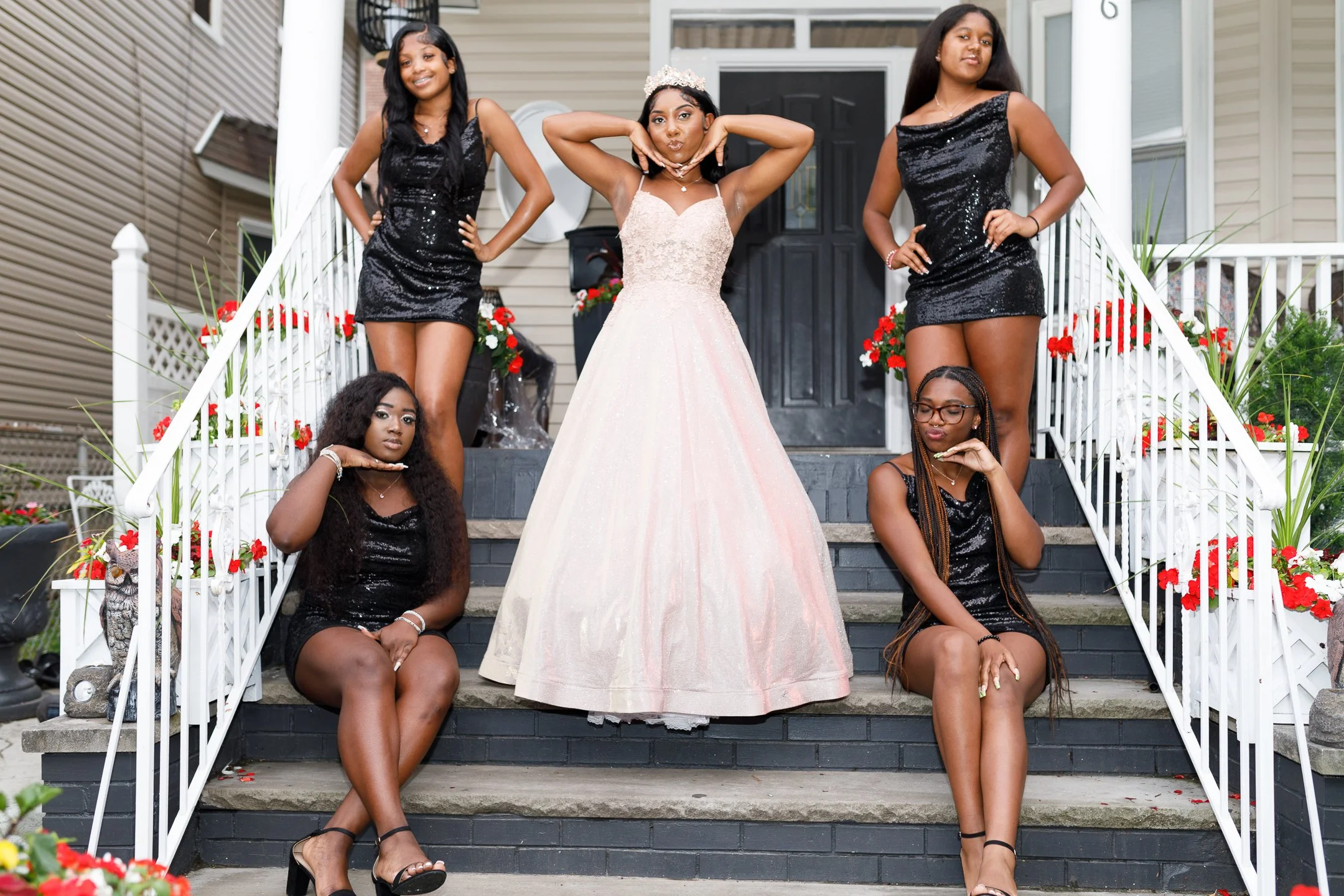 Six women, one in a formal gown and the others in black dresses, are posing on a front porch decorated with red and white flowers. The woman in the gown is standing at the center, surrounded by the other women who are either sitting or standing on th