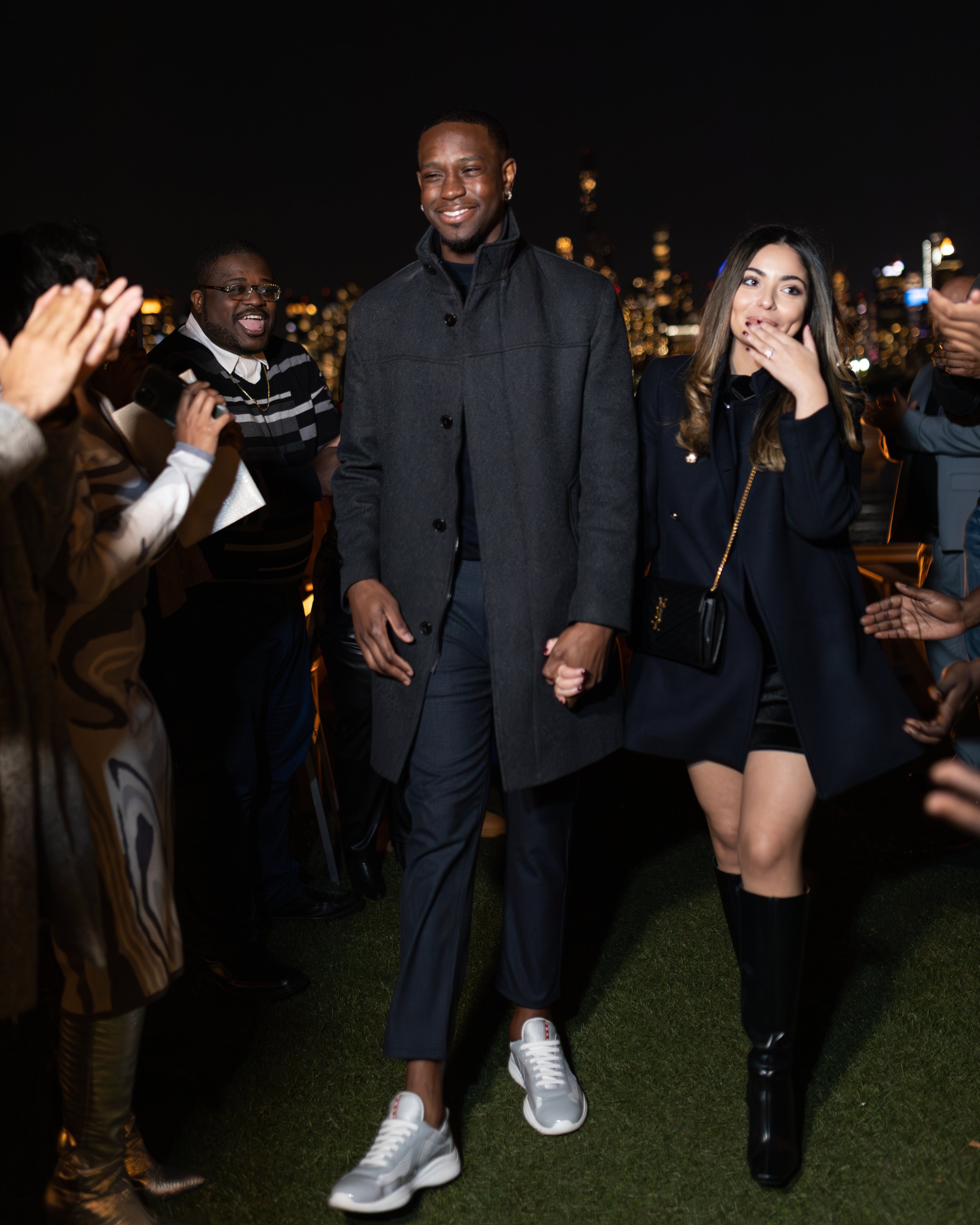A man and woman holding hands at night, surrounded by friends in a city skyline background.