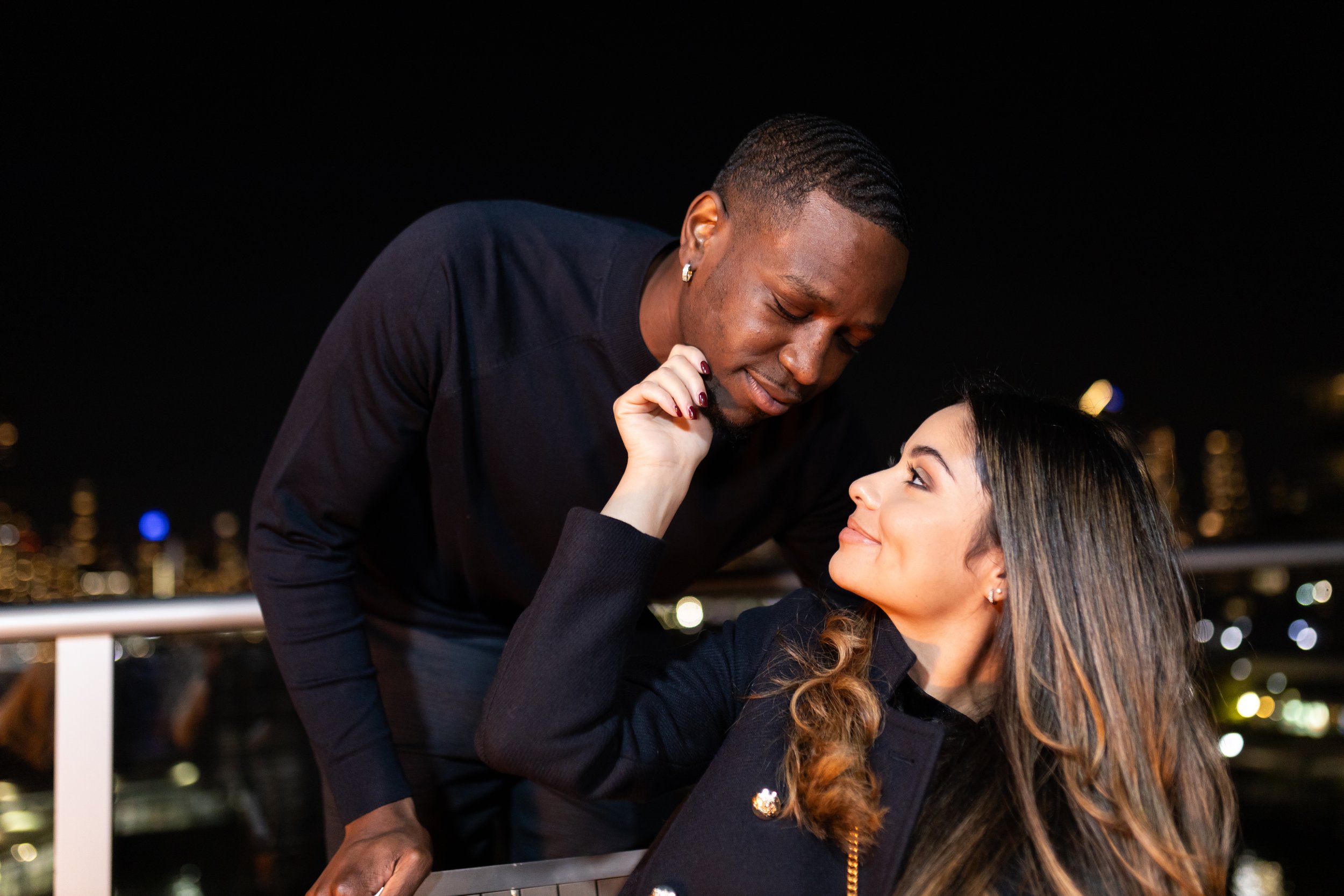A man leaning over a woman at night, close to her face, with blurred city lights in the background. The woman is smiling and gazing up at the man.