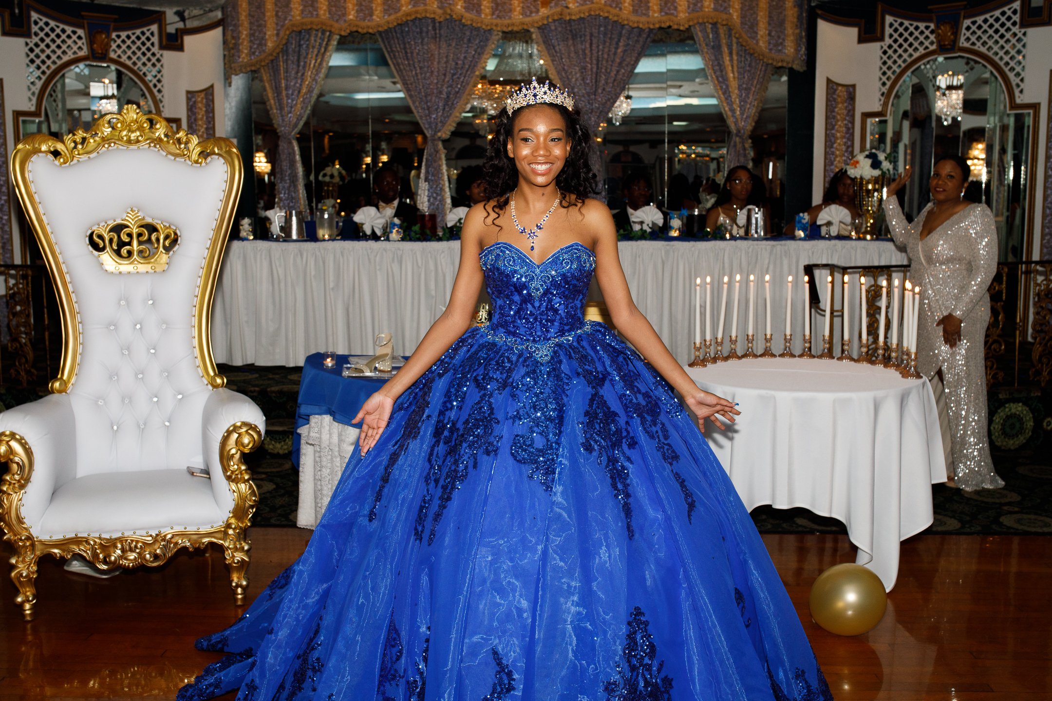 A young woman in a royal blue, beaded quinceañera dress with a tiara and necklace, standing in a decorated banquet hall with a princess throne chair and a table with candles in the background.