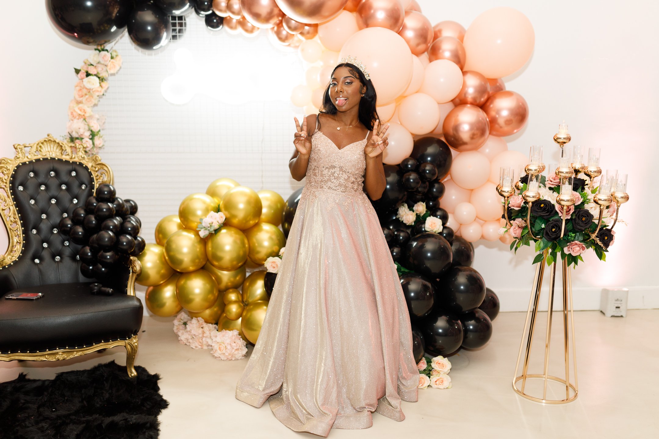A girl in a fancy gown making a peace sign and a playful face, standing in front of a backdrop of pink, black, and gold balloons and a floral arrangement with dark roses, at a celebration or party.