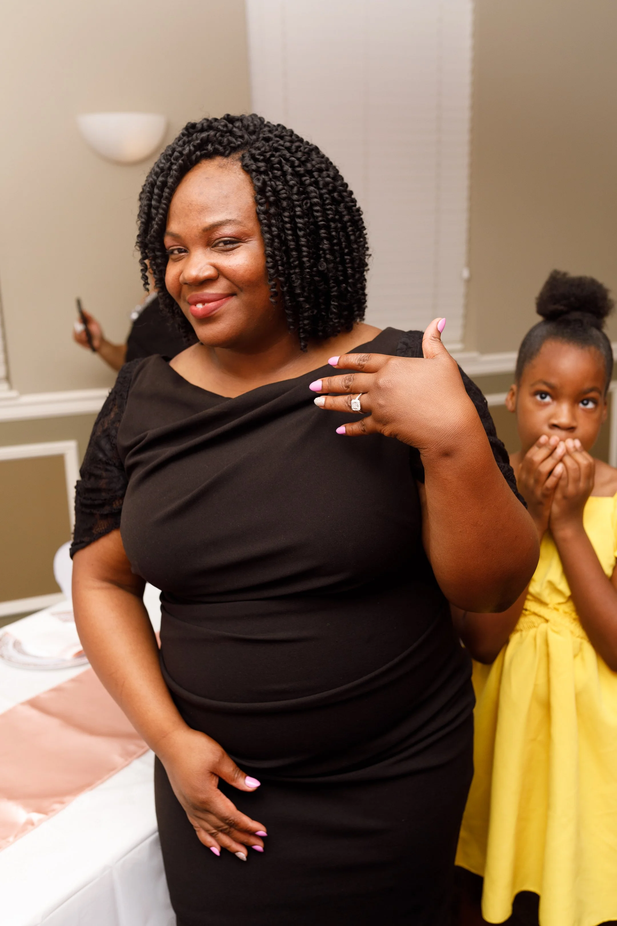 A woman with curly black hair in a black dress, smiling and showing off an engagement ring, with a young girl in a yellow dress covering her mouth in the background.