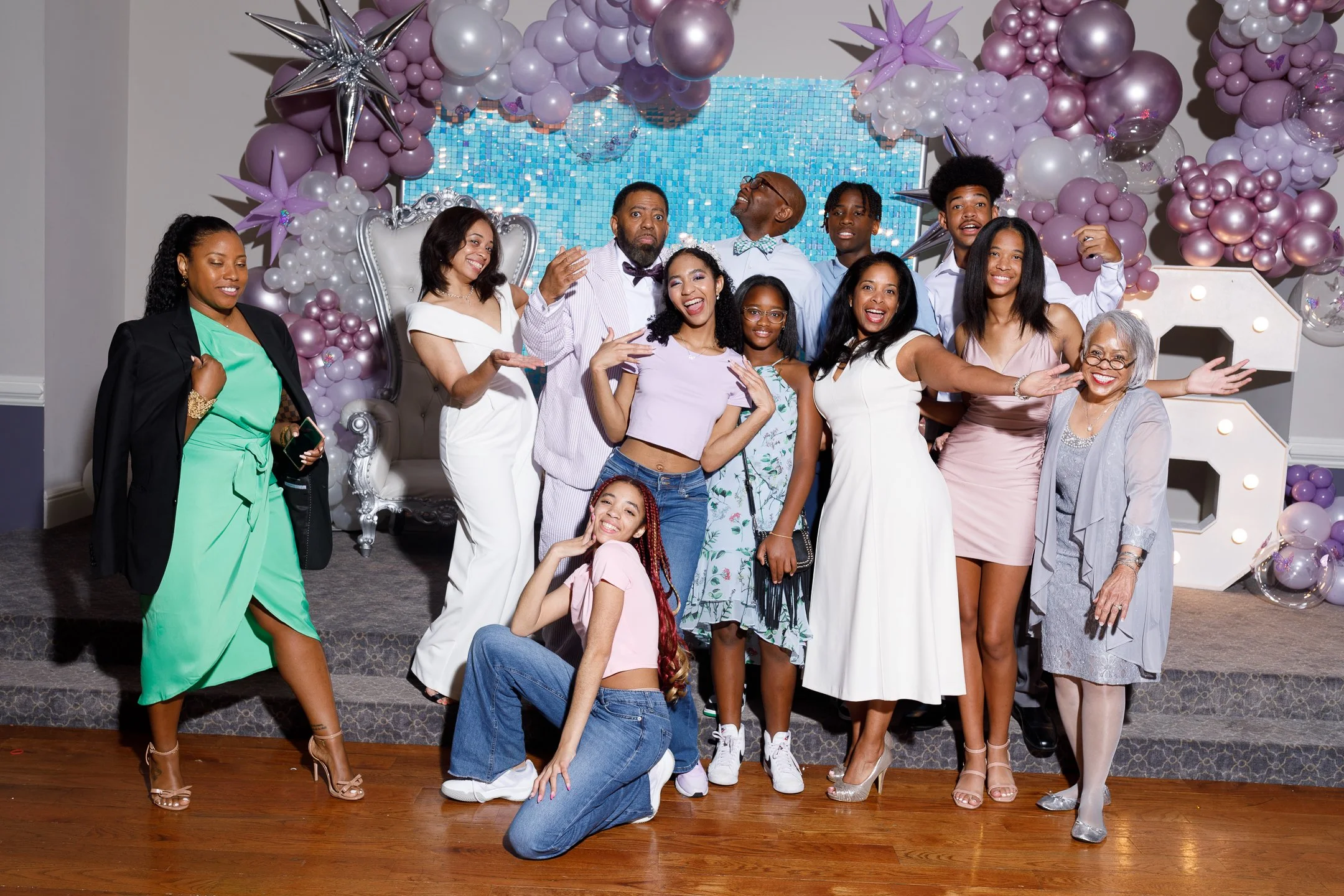 Group of people celebrating at a party with pastel pink and purple balloons, a decorated backdrop, and large illuminated number '3'.