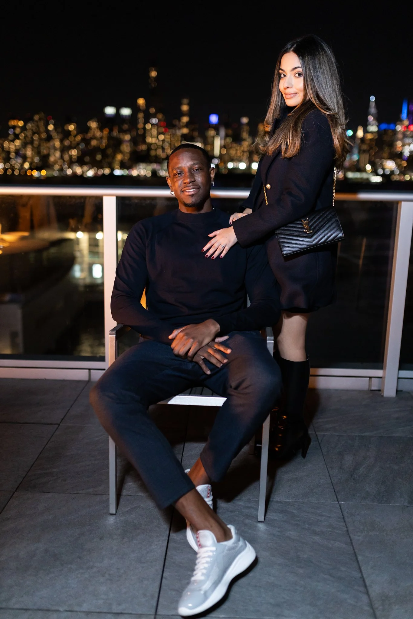 A man and a woman on a rooftop at night with a city skyline in the background. The man is sitting on a chair, and the woman is standing next to him with her hand on his shoulder. They are both dressed stylishly, and the woman is wearing a black outfi