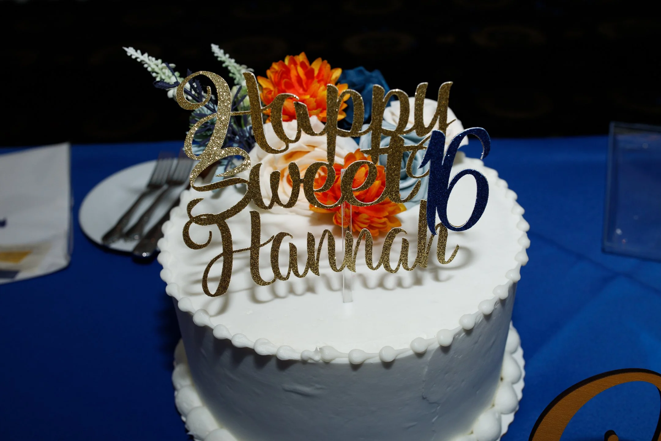 A white frosted cake decorated with orange flowers and greenery, topped with a gold glittery cake topper that reads 'Happy Sweet 16 Hannah', on a blue tablecloth.