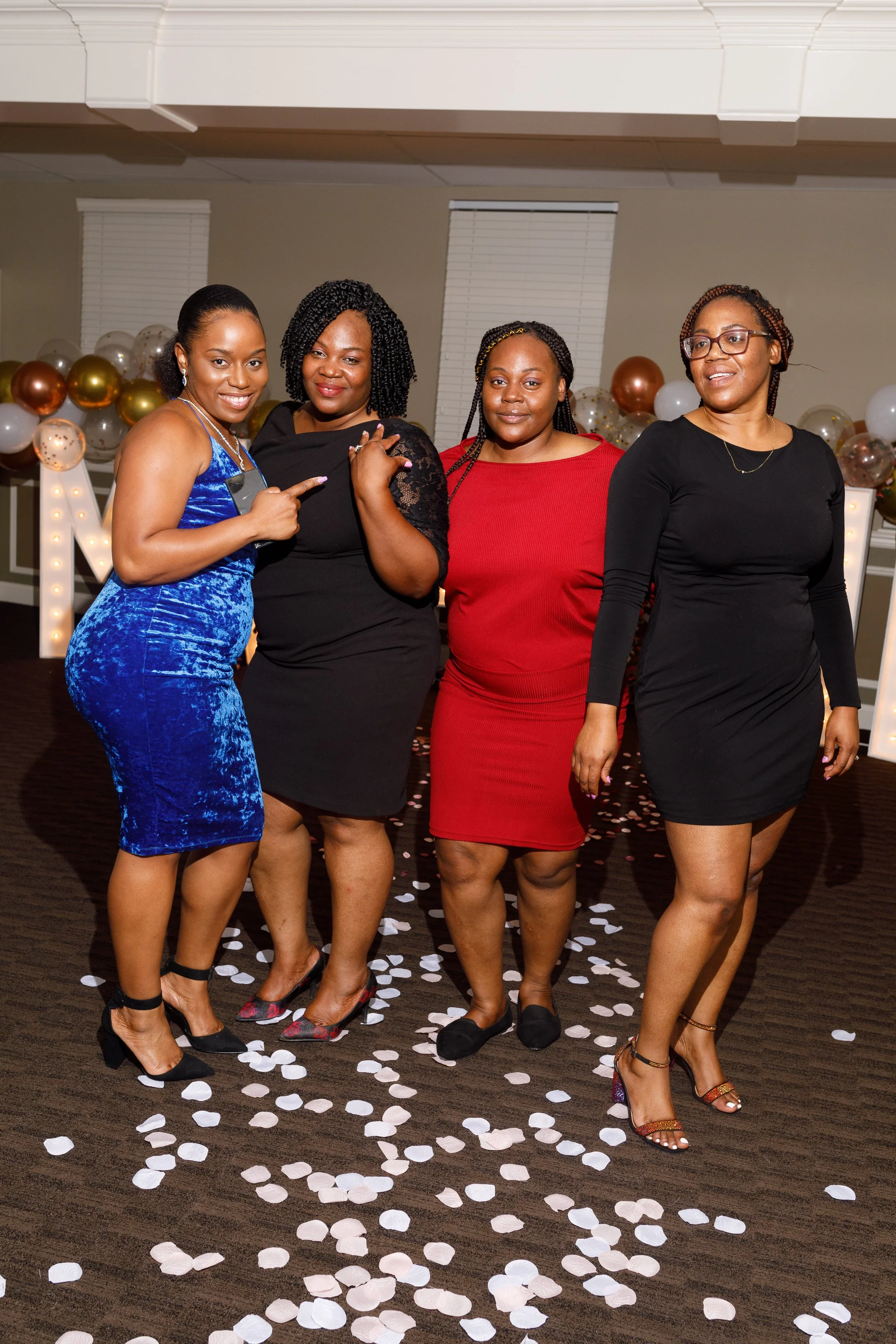 Four women dressed formally, standing on a decorated floor with white confetti, at a celebration or party.