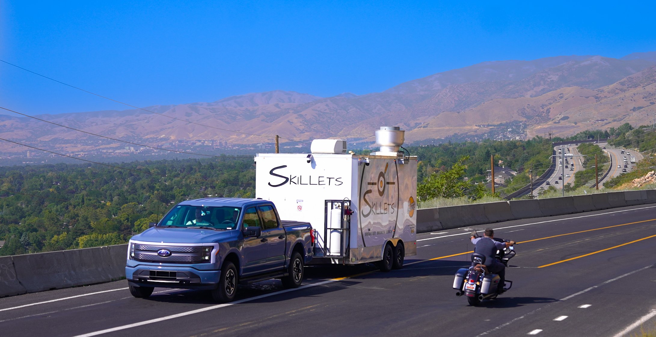 A blue pickup truck towing a trailer that reads "Skillets" on a highway near a green landscape and distant mountains under a clear blue sky.