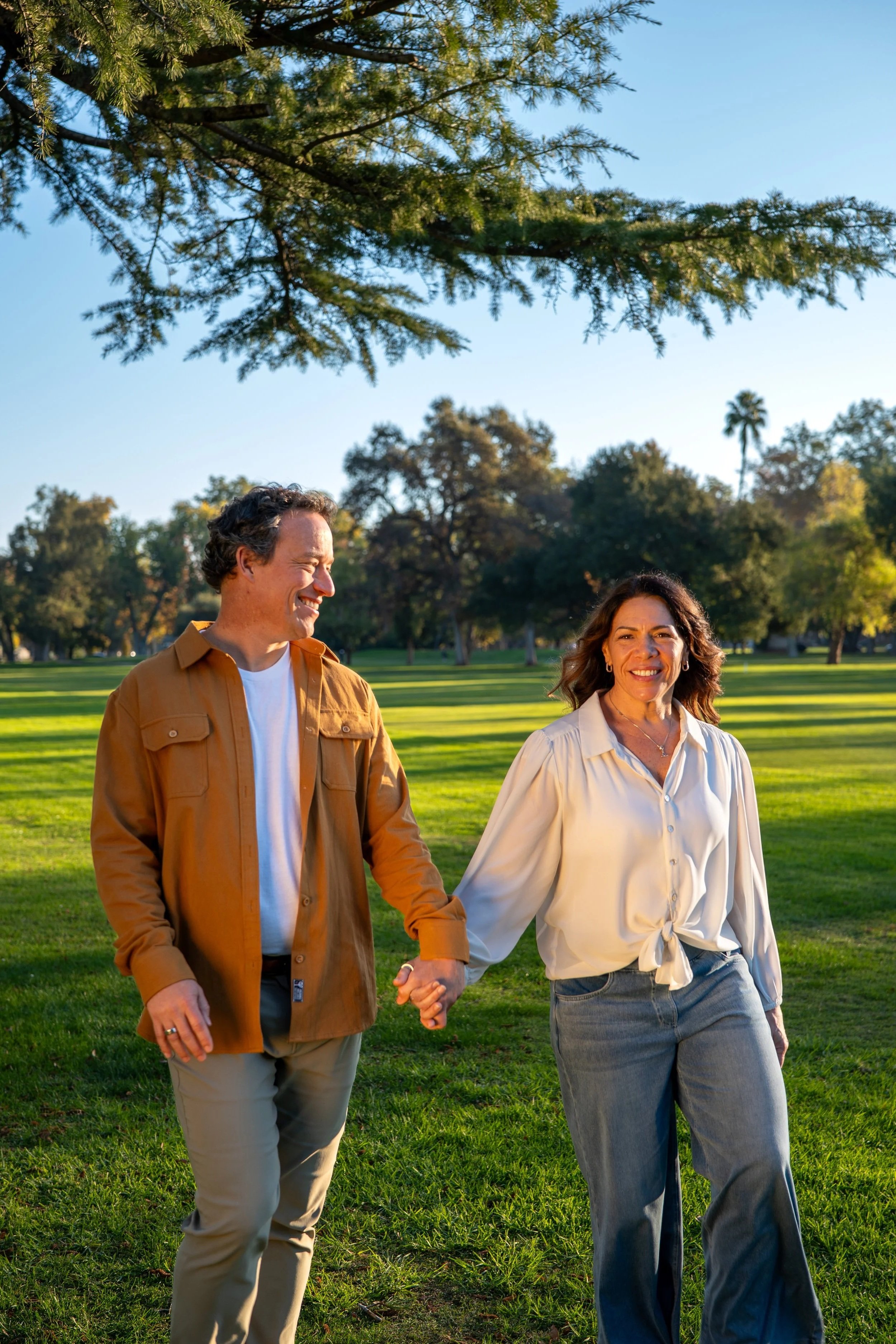 A couple walking hand in hand in a park, smiling and enjoying a sunny day with trees and a clear blue sky in the background.