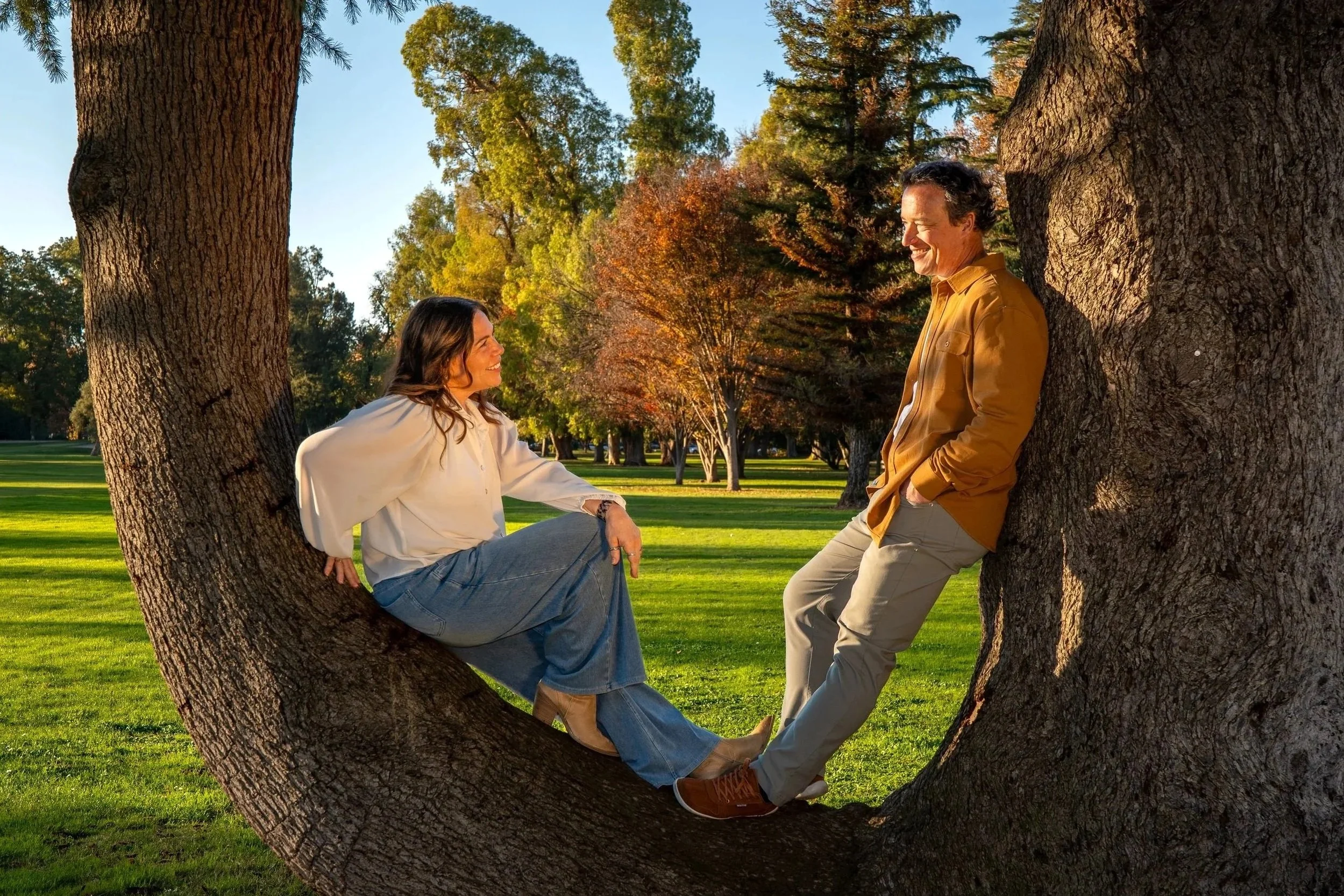 A woman and a man are smiling and looking at each other while sitting and leaning against a large tree in a park with green grass and colorful autumn trees.