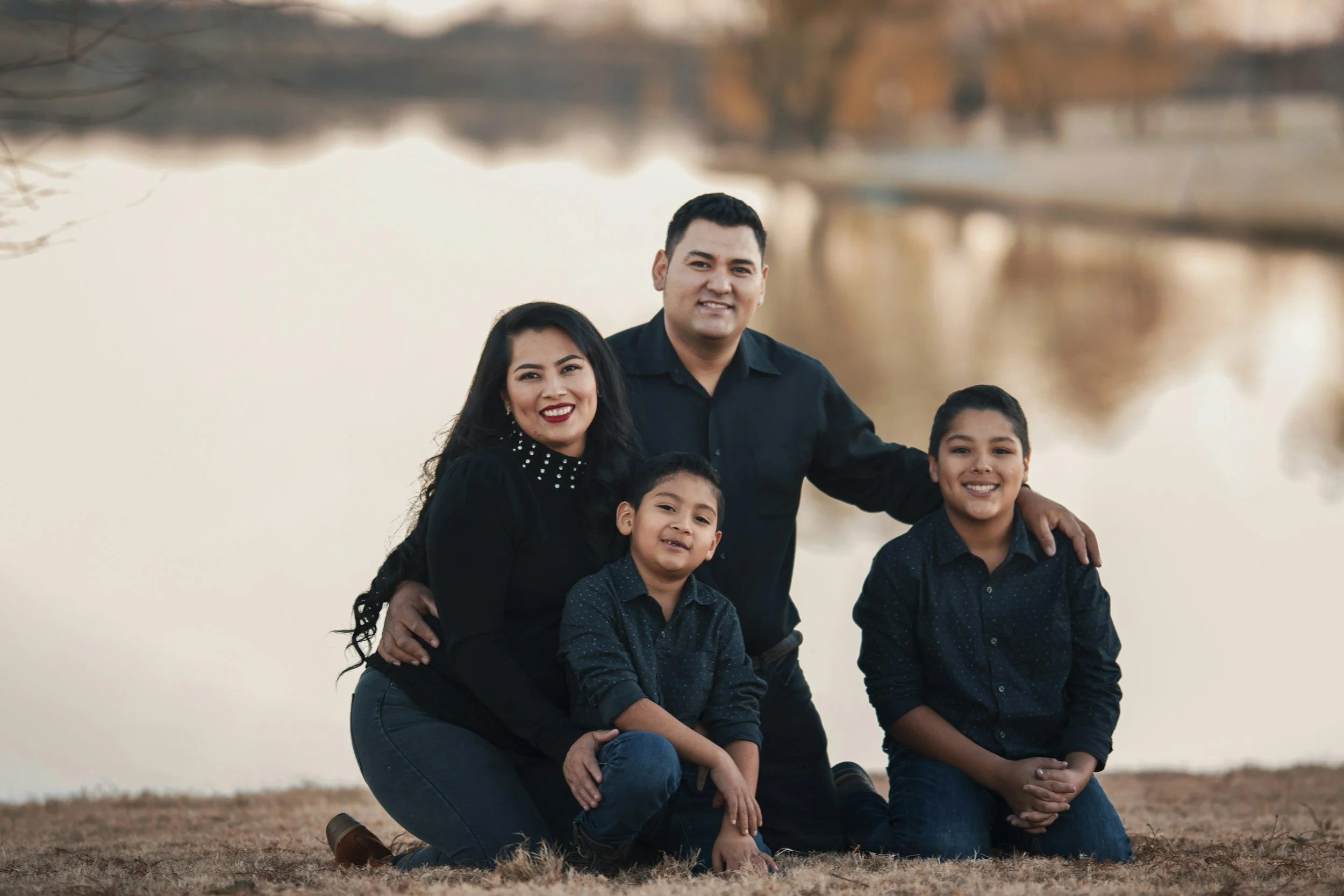 A family of four posing outdoors near a body of water during sunset, smiling at the camera.