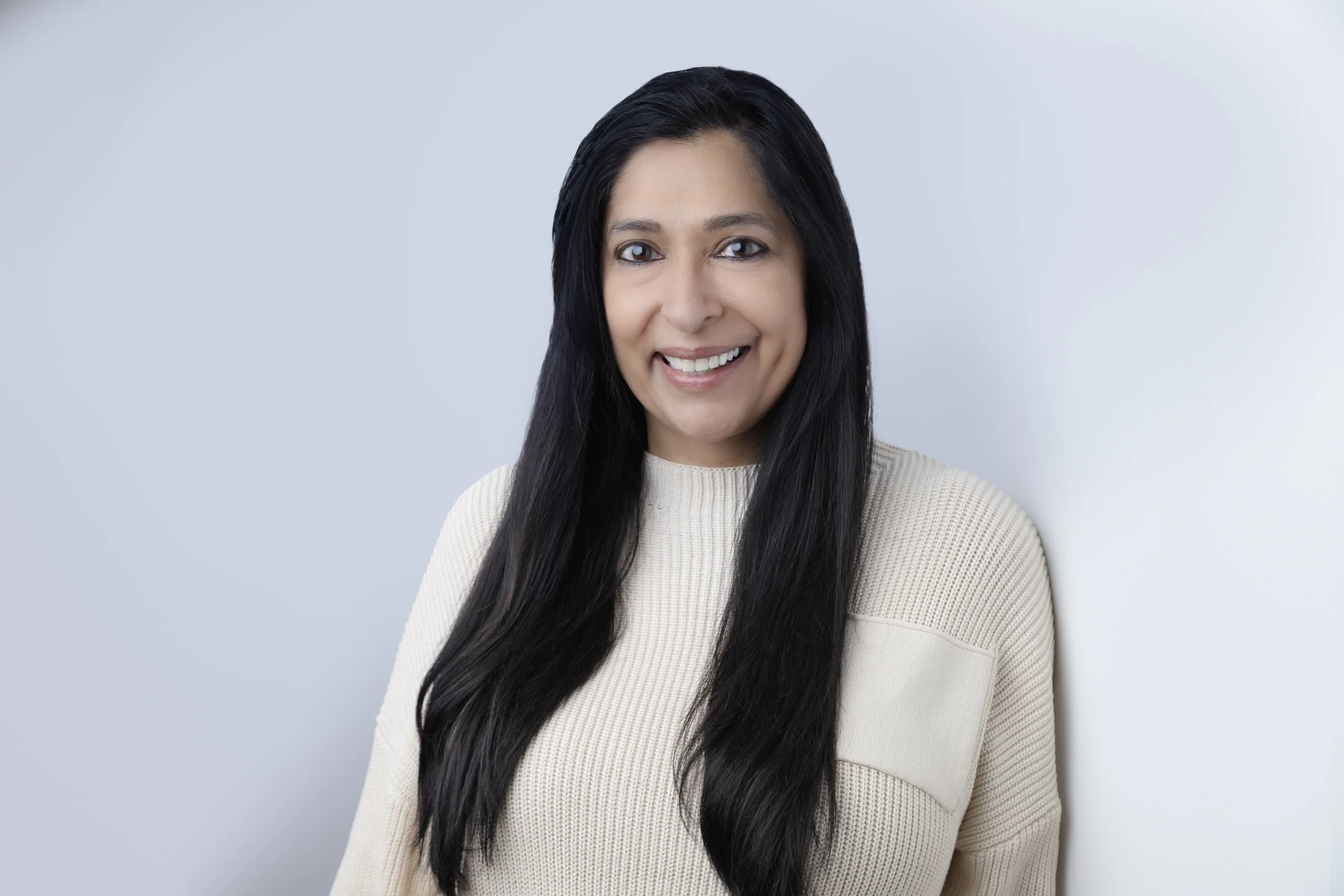 A woman with long black hair smiling, wearing a cream-colored sweater, standing against a light gray background.