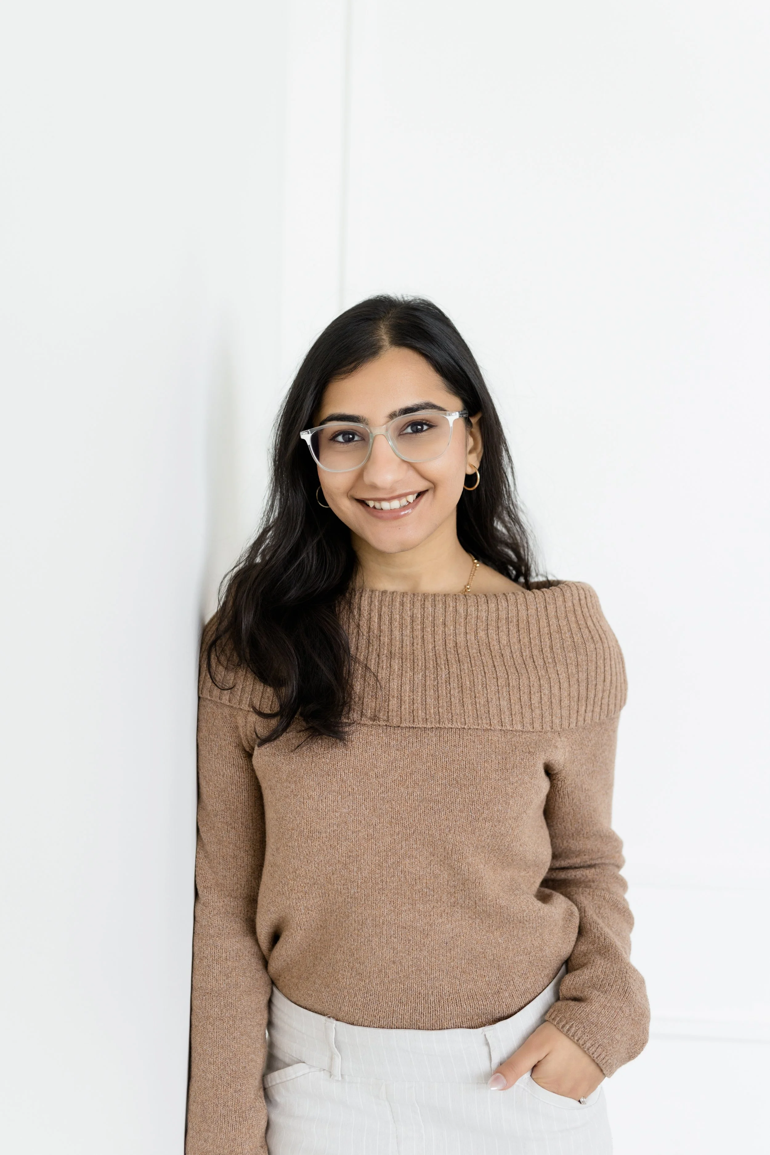 A woman with long dark hair, glasses, and hoop earrings smiling while standing against a white background, wearing a tan sweater and white pants.
