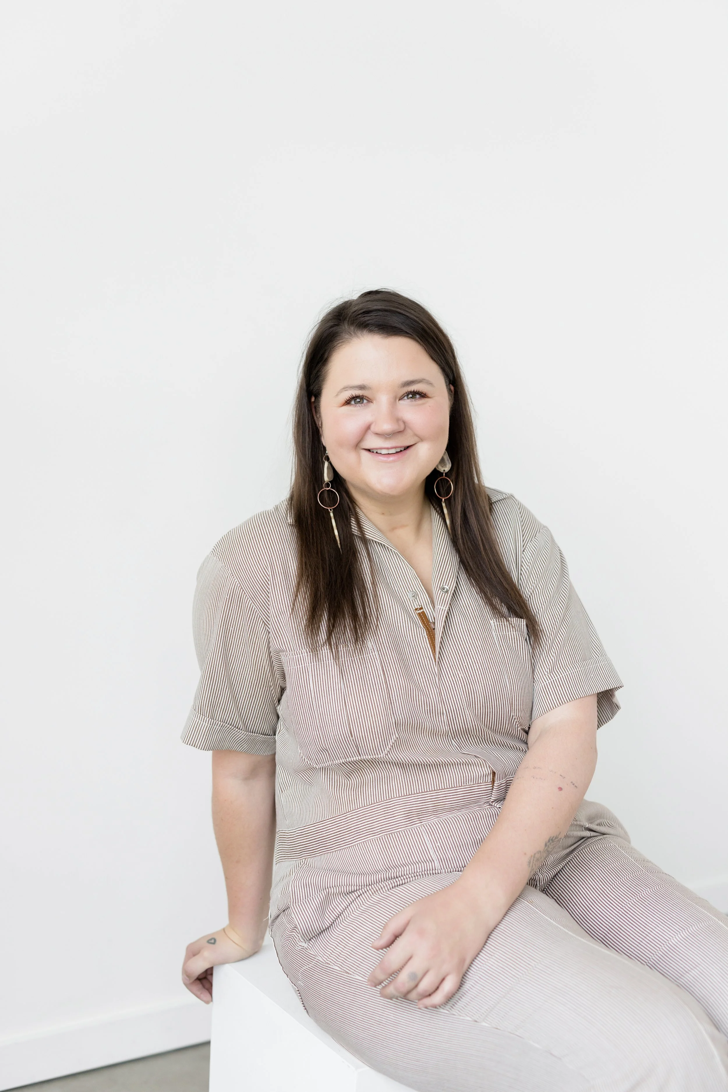 A woman with long brown hair, wearing a beige striped shirt and earrings, sitting on a white block against a white wall, smiling at the camera.
