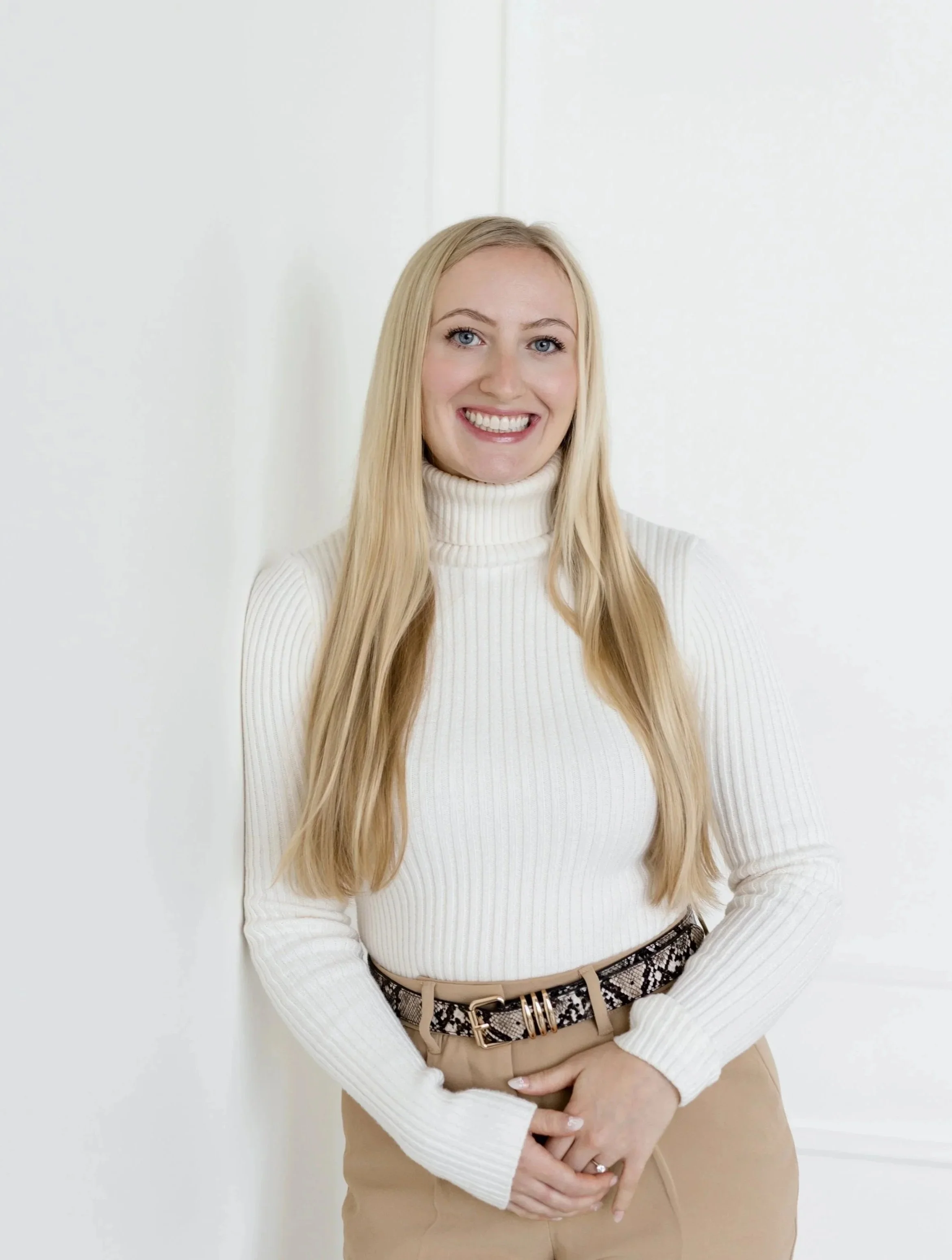 A young woman with long blonde hair wearing a white turtleneck sweater and beige pants, standing against a white wall, smiling.