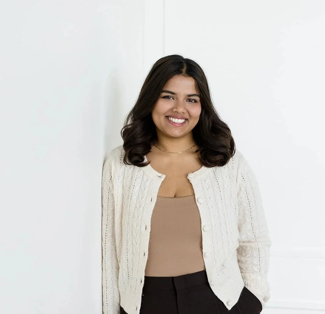 Young woman with shoulder-length brown hair and a warm smile, wearing a cream-colored cardigan and a white top, posing against a plain beige background.