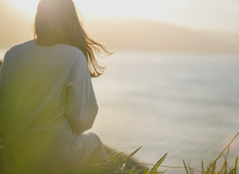 A woman sitting near a body of water during sunset or sunrise, with her back facing the camera and her hair blowing in the wind.