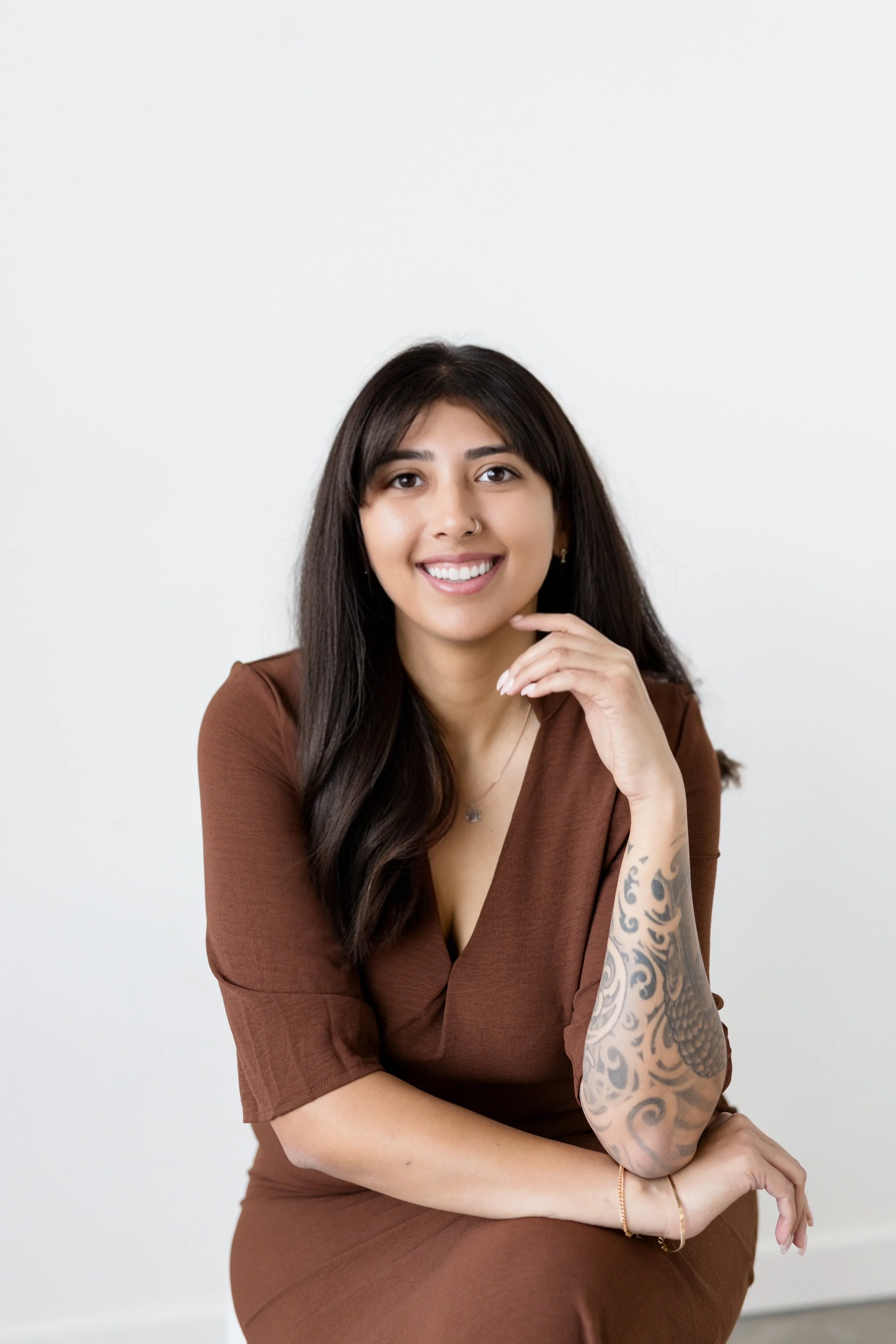 A young woman with long dark hair, wearing a brown dress, smiling and sitting against a plain white background.