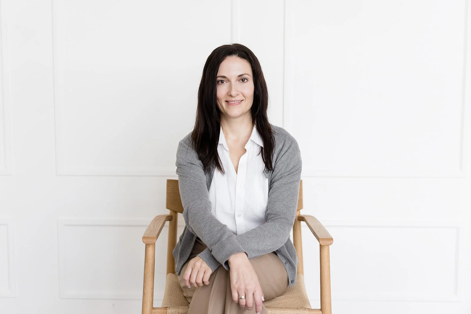 A woman with long dark wavy hair, wearing a beige blazer over a light-colored blouse, sitting on a light-colored couch with an orange pillow, against a plain white wall.