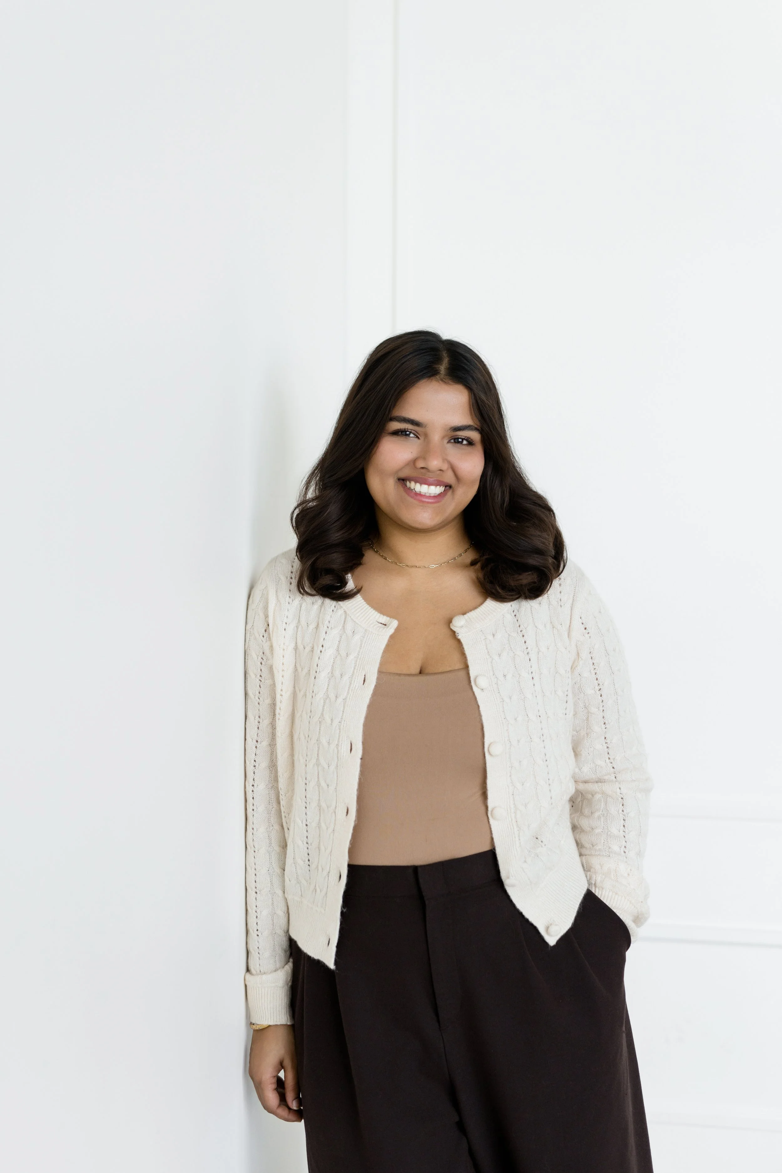 A young woman with dark hair, wearing a beige top, a cream-colored cardigan, and dark pants, standing against a plain white background, smiling.