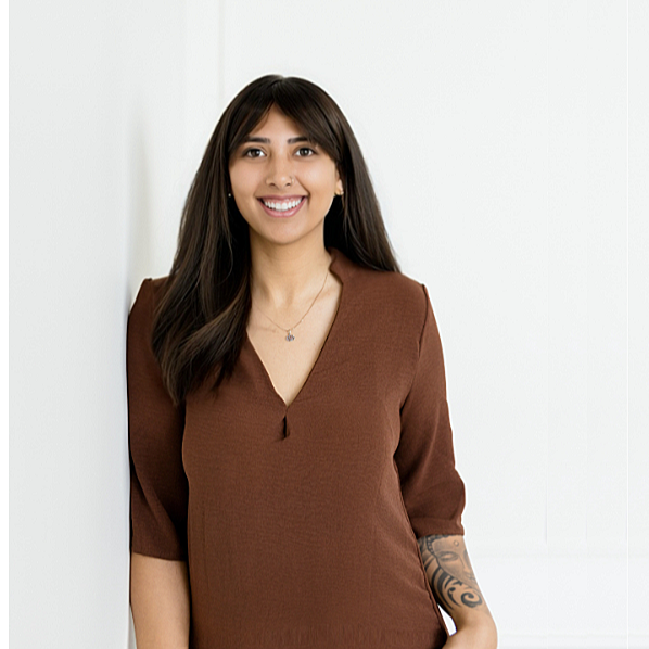 Young woman with long dark hair smiling, wearing a brown blouse, standing indoors against a white wall.