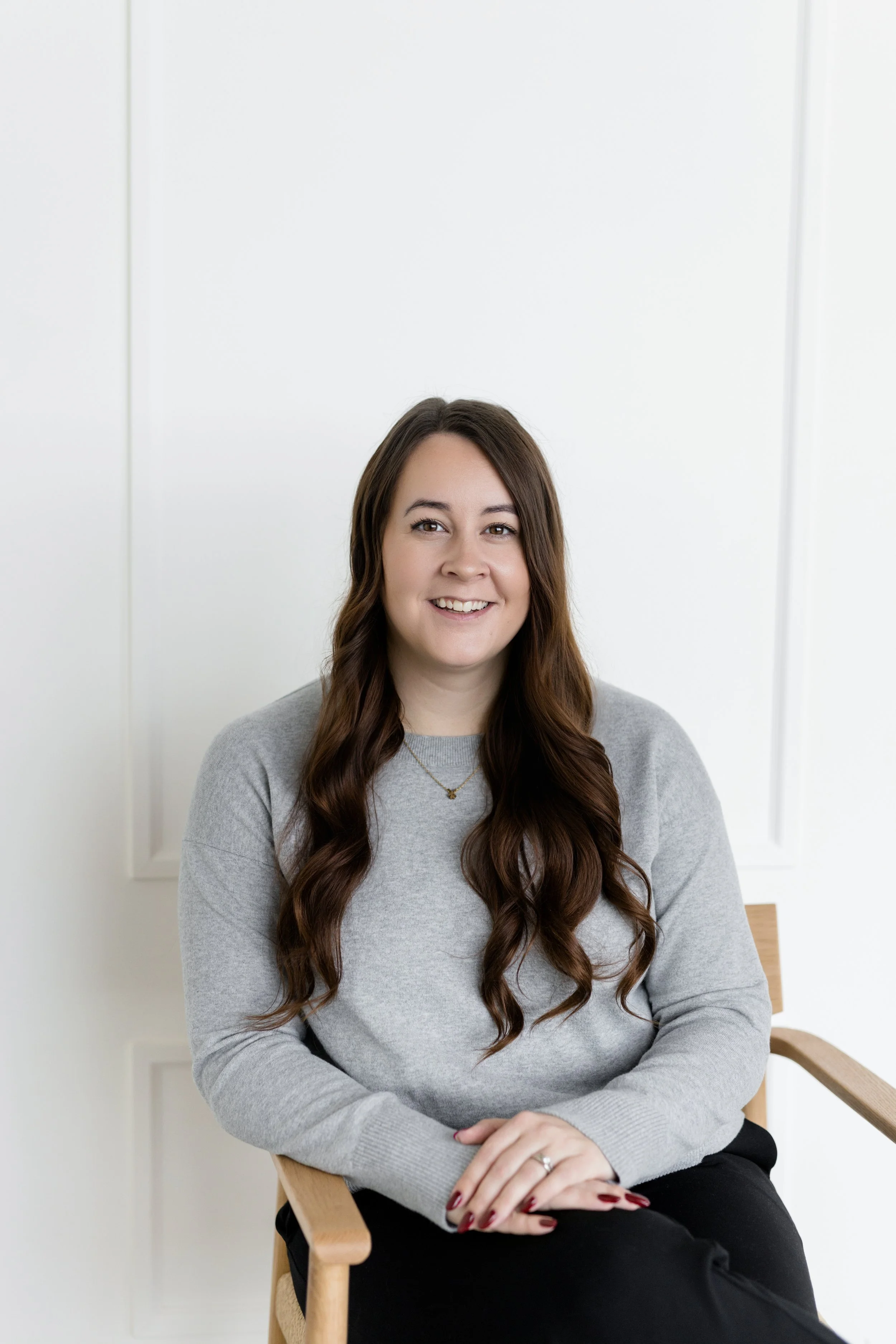 A woman with long brown hair in loose waves, wearing a light gray sweater and black pants, sitting on a wooden chair with a light wood armrest, smiling, inside a bright room with white walls and minimal decor.