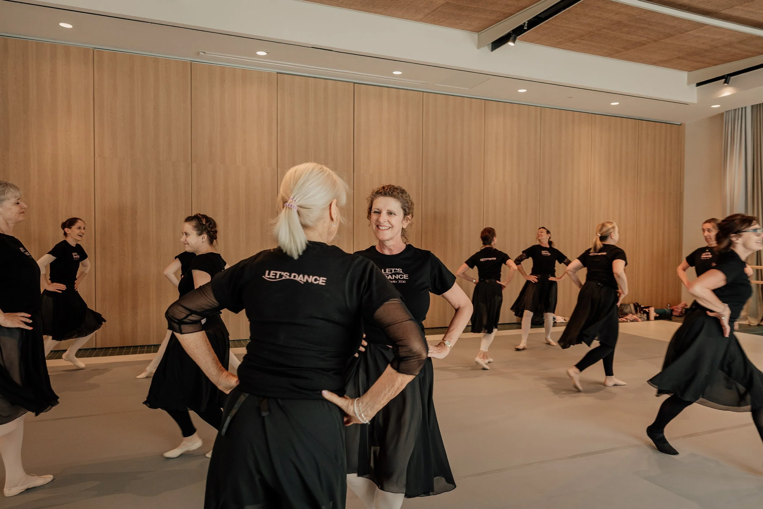 Group of women practicing a synchronized dance routine in a Spanish studio, wearing black "Let's Dance" shirts and skirts.