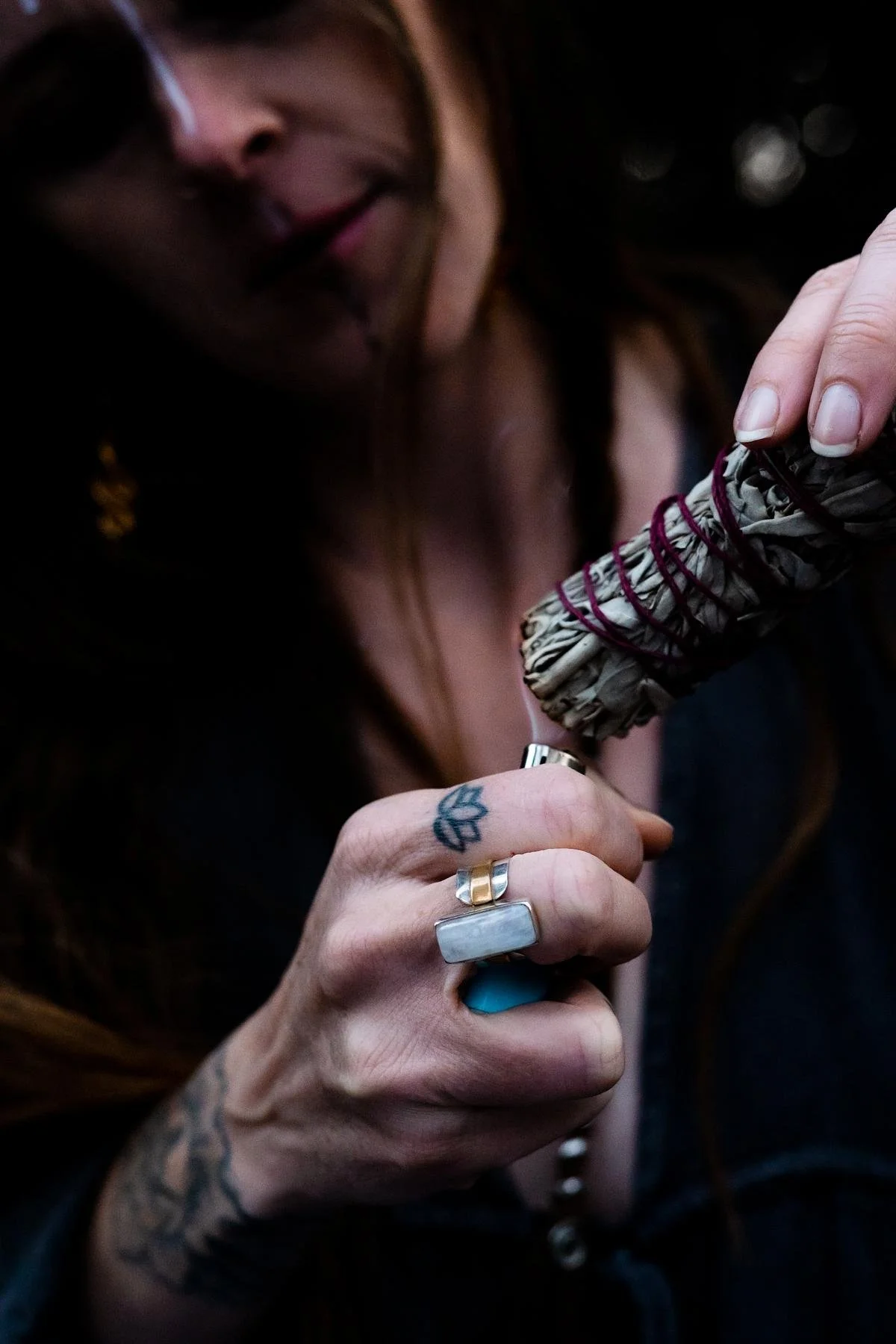 Women wearing blue rings with a lotus tattoo is burning a bundle of sage with a lighter.
