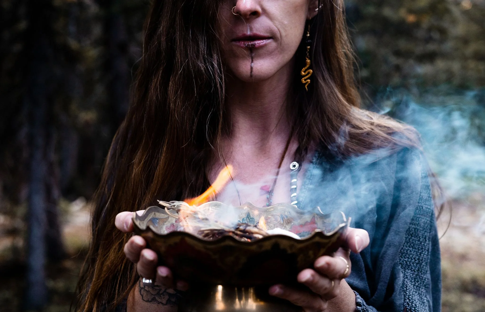 Shamanic woman with face paint and snake earrings holds a bowl with both hands. The bowl has sage burning causing smoke and fire.