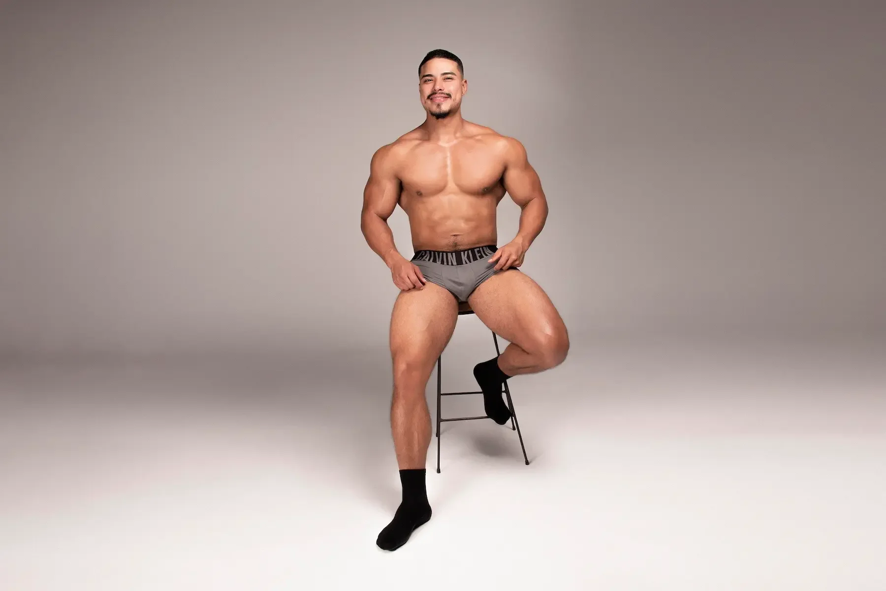 Hombre musculoso sentado en una silla, usando solo calzoncillos grises y calcetines negros, posando con una sonrisa y fondo gris liso.