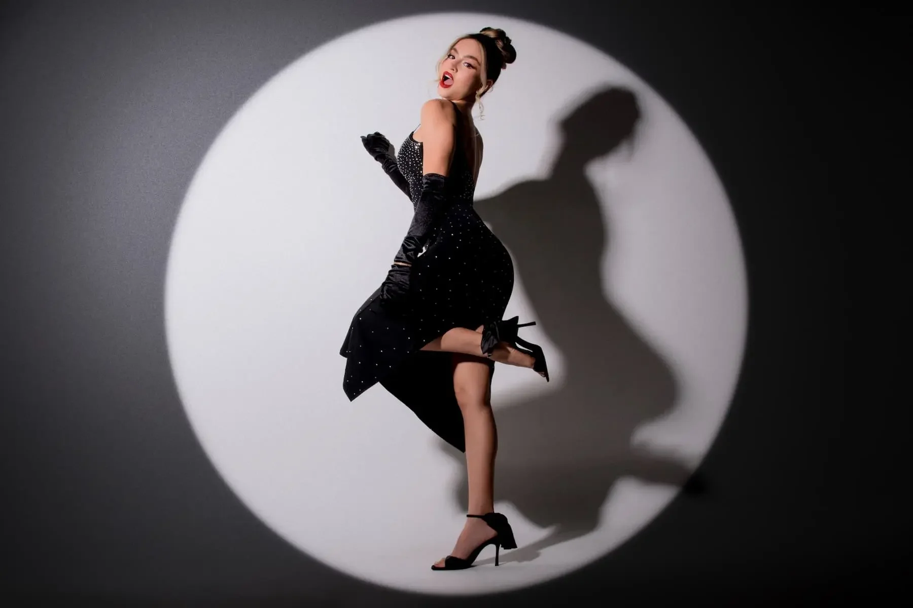 Mujer en vestido negro con lunares blancos, con guantes largos y tacones, posando con expresión atrevida y levantando una pierna, en un fondo de luz que crea sombra en la pared.