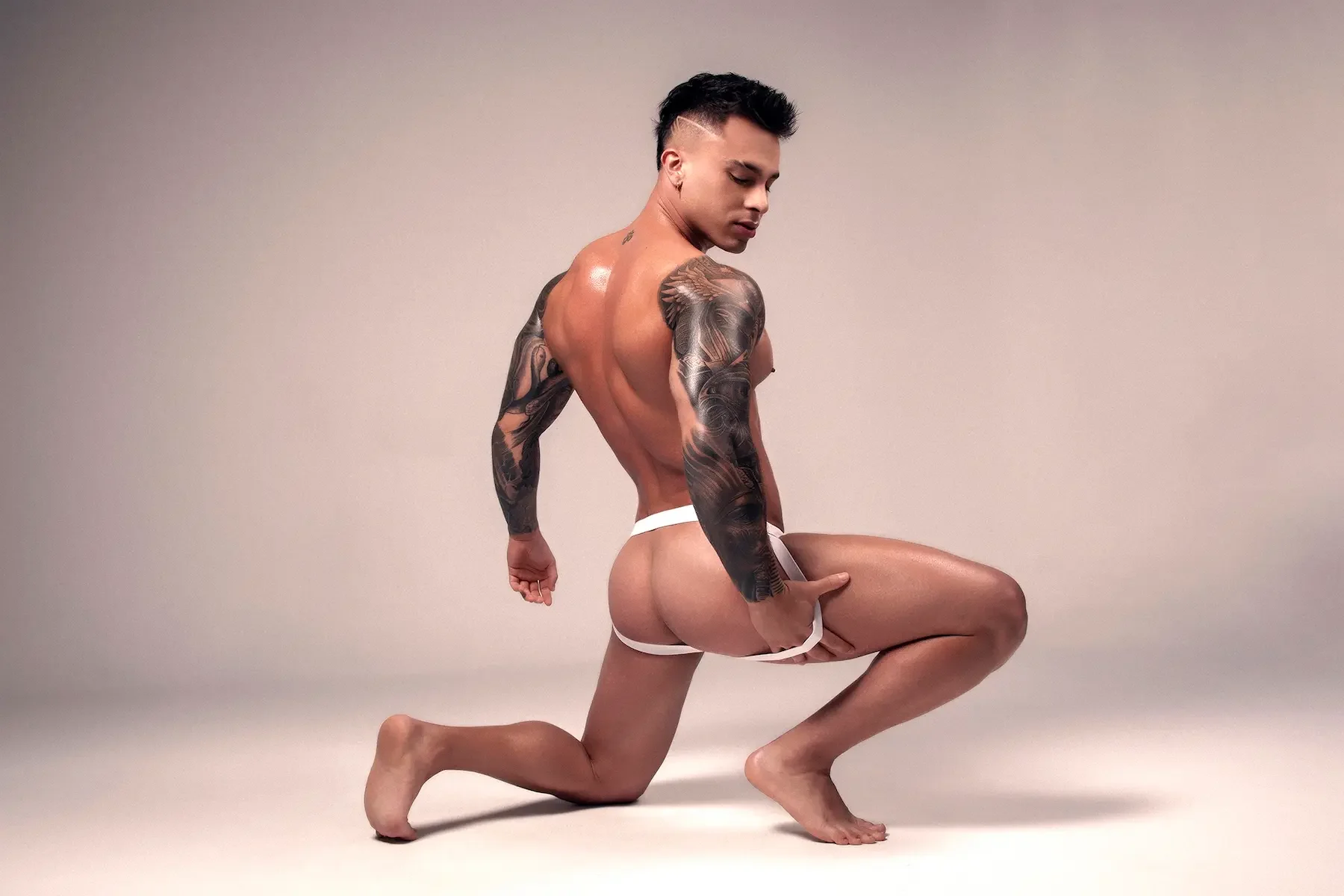 Un hombre musculoso con tatuajes en brazos y espalda se arrodilla en el suelo, usando solo ropa interior blanca, en un fondo neutro.