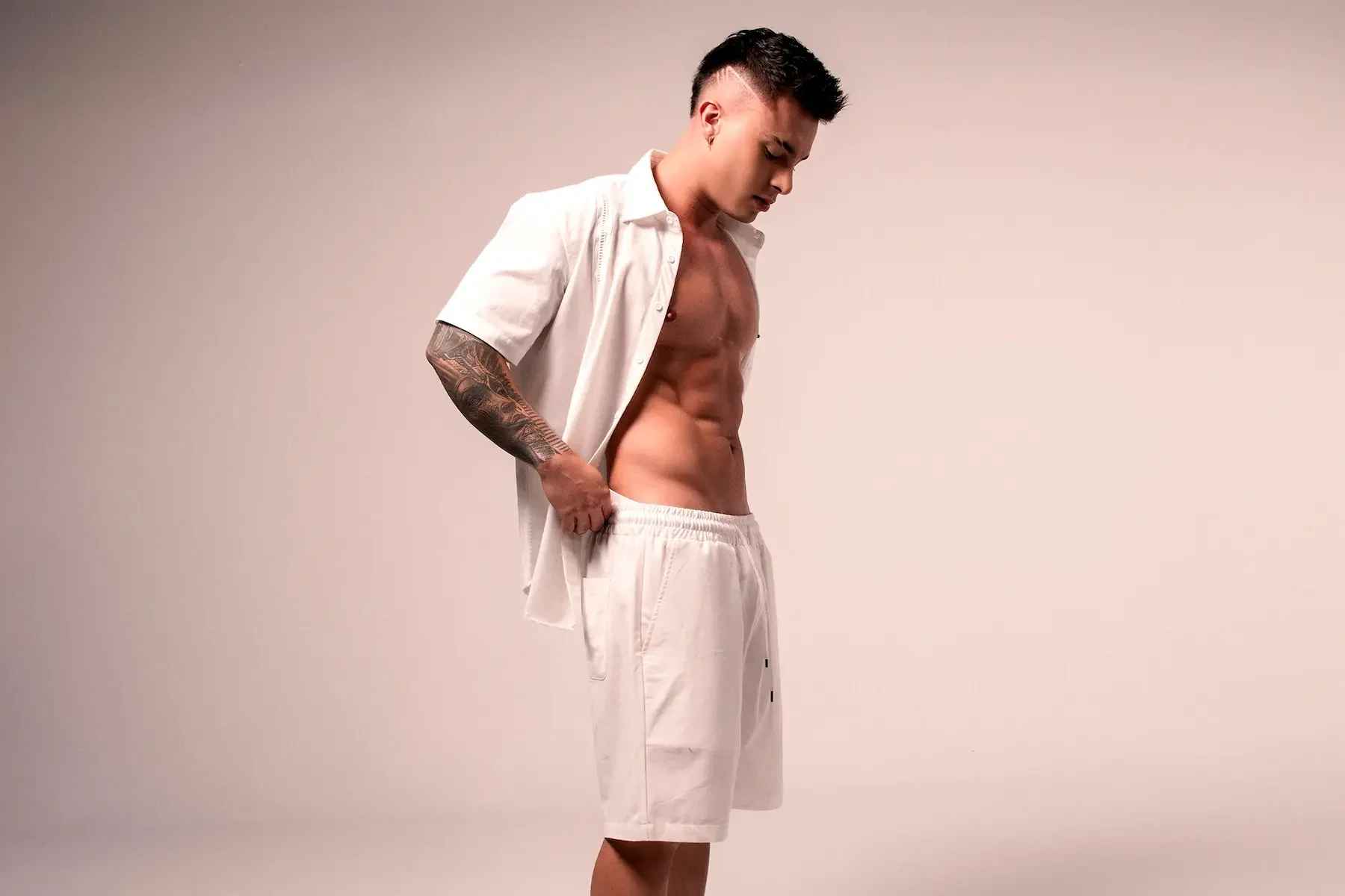 Joven con peinado de punta, sin camisa y con tatuajes en el brazo, ropa blanca shorts y camisa abierta, mirando hacia abajo, en un fondo neutro.