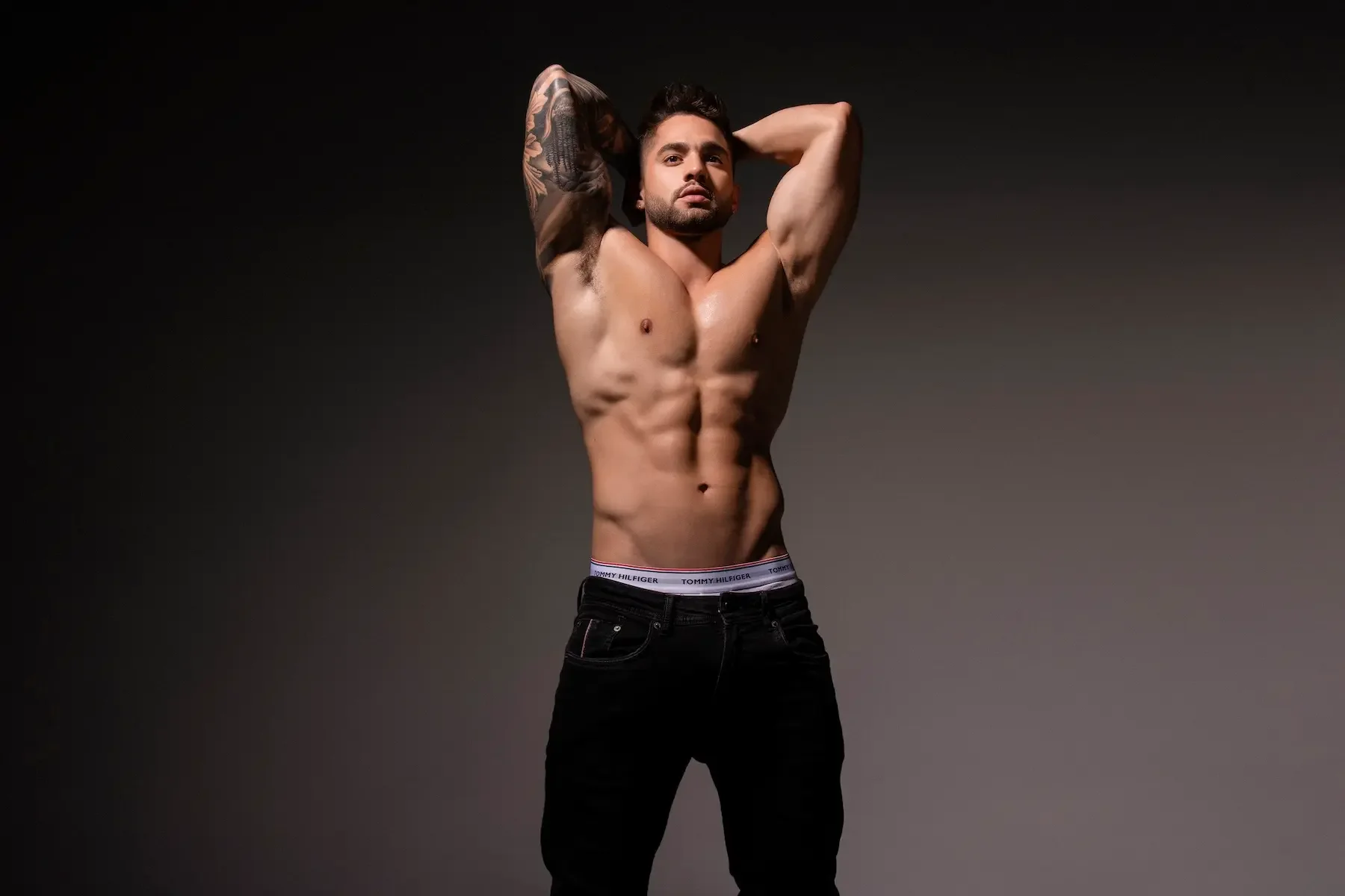 Joven hombre con torso desnudo y pantalones negros, con un tatuaje en su brazo izquierdo, posando con las manos detrás de la cabeza, en un fondo oscuro.