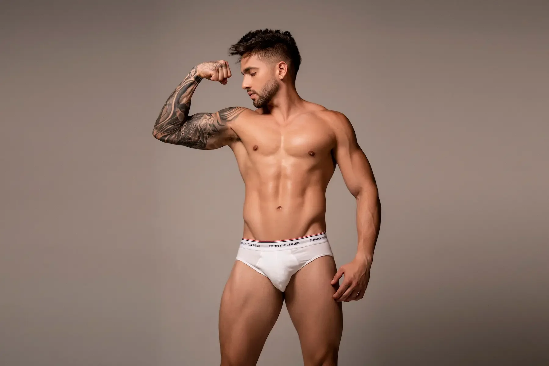 Un hombre joven sin camisa con tatuajes en el brazo izquierdo, vistiendo ropa interior blanca de la marca Tommy Hilfiger, flexionando el brazo izquierdo y mirando hacia abajo, en un fondo gris.
