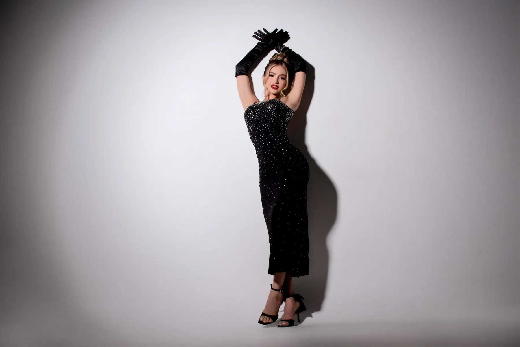 Mujer con vestido negro con puntos blancos, guantes negros largos, tacones negros, posando con brazos elevados y cruzados sobre su cabeza, en un fondo gris con sombra.