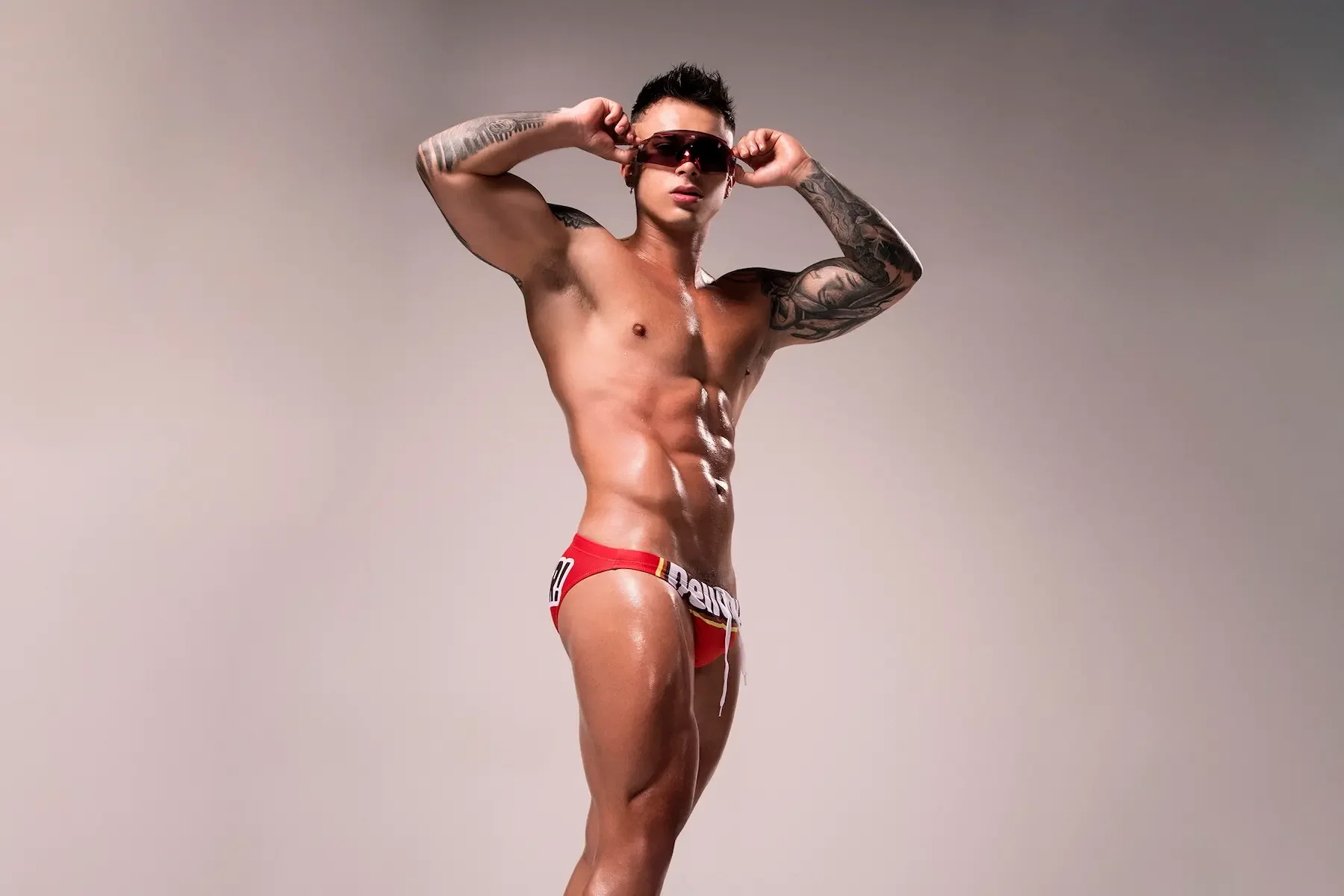 Joven con tatuajes ajustándose gafas de sol, vistiendo ropa interior roja, en un fondo neutro.