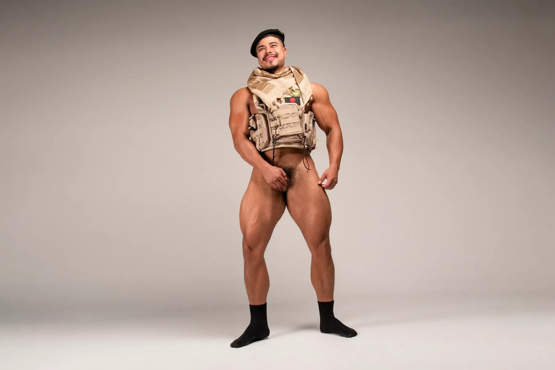 Hombre con mochila militar, sin ropa, en un fondo gris, sonriendo, usando calcetines negros.